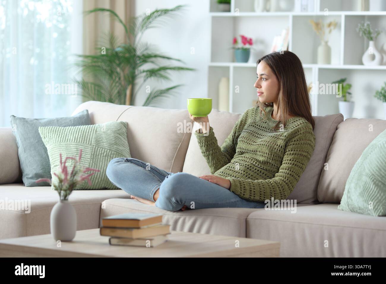 Frau in Grün, die Kaffee trinkt, sitzt auf einer Couch in einem Haus drinnen Stockfoto