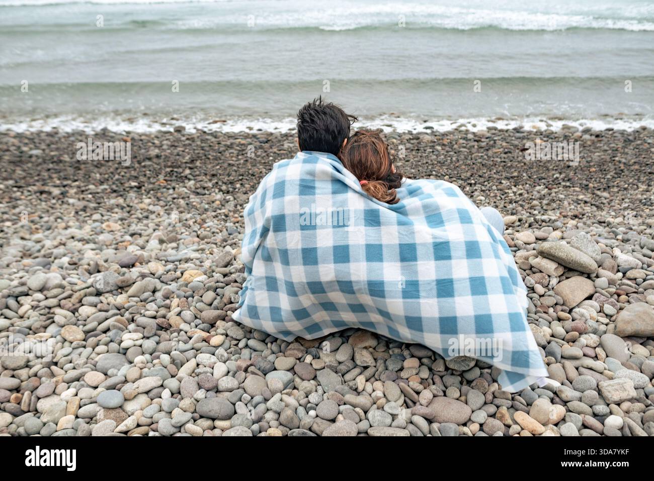 Ein Paar sitzt zusammen unter einer Decke an einem felsigen Strand und blickt auf das Meer und den bewölkten Winterhimmel. Stockfoto
