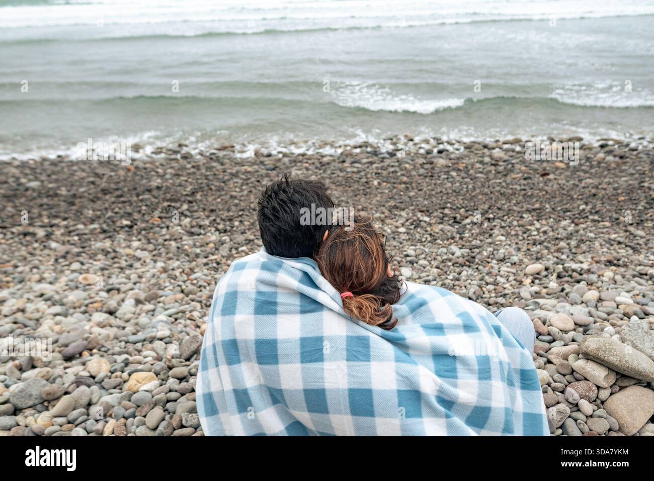 Ein Paar sitzt zusammen unter einer Decke an einem felsigen Strand und blickt auf das Meer und den bewölkten Winterhimmel. Stockfoto