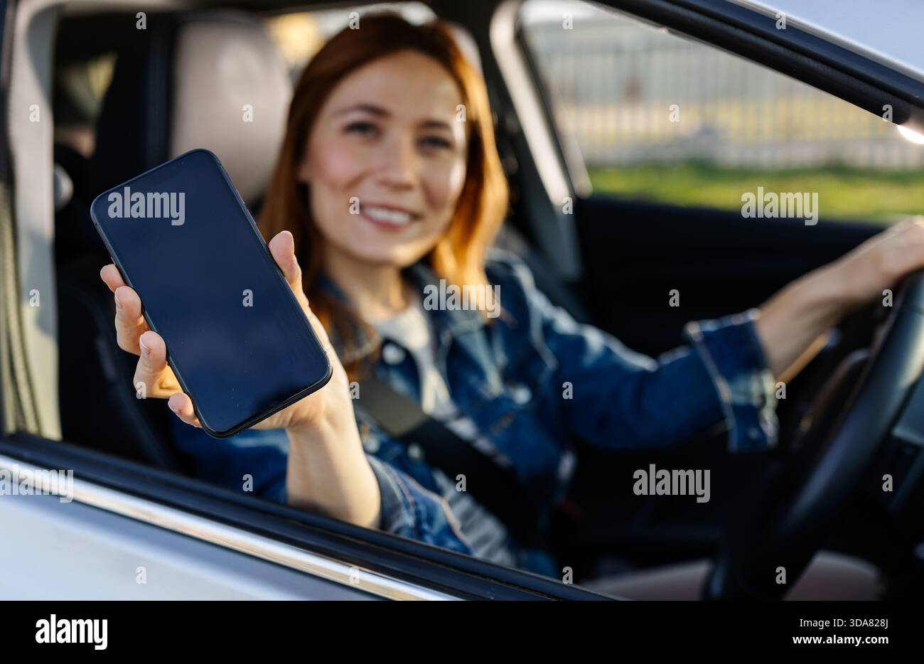 Eine Frau sitzt in einem geparkten Auto und hält ihr Smartphone mit einem Lächeln zur Kamera. Es ist Tag und sie scheint glücklich zu sein. Stockfoto