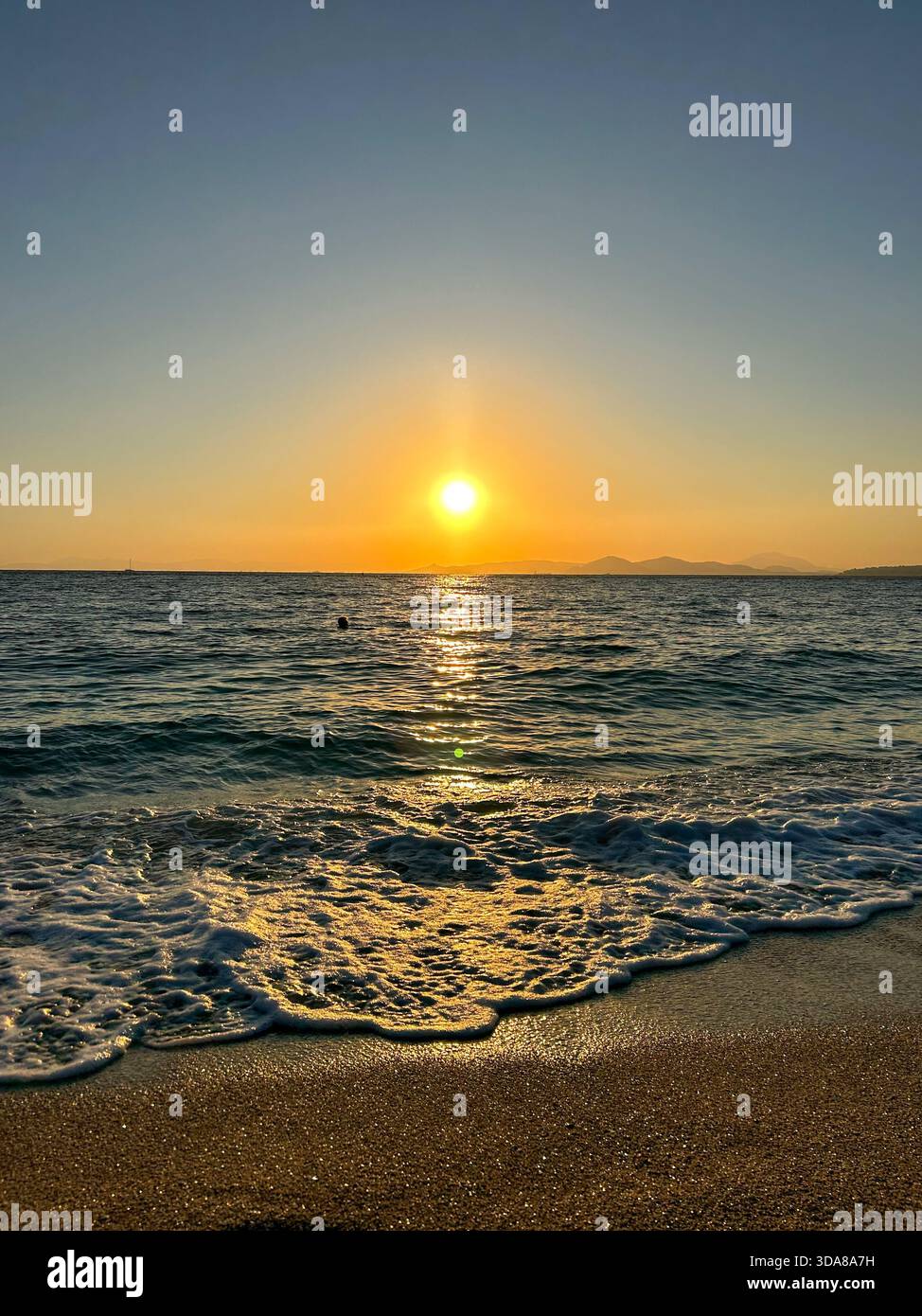 Ruhige Wellen, die bei einem goldenen Sonnenuntergang auf einen Sandstrand gleiten, während die Sonne über dem Meer reflektiert wird und entfernte Silhouetten der Berge. - Smartphone-aufgenommenes Stockfoto