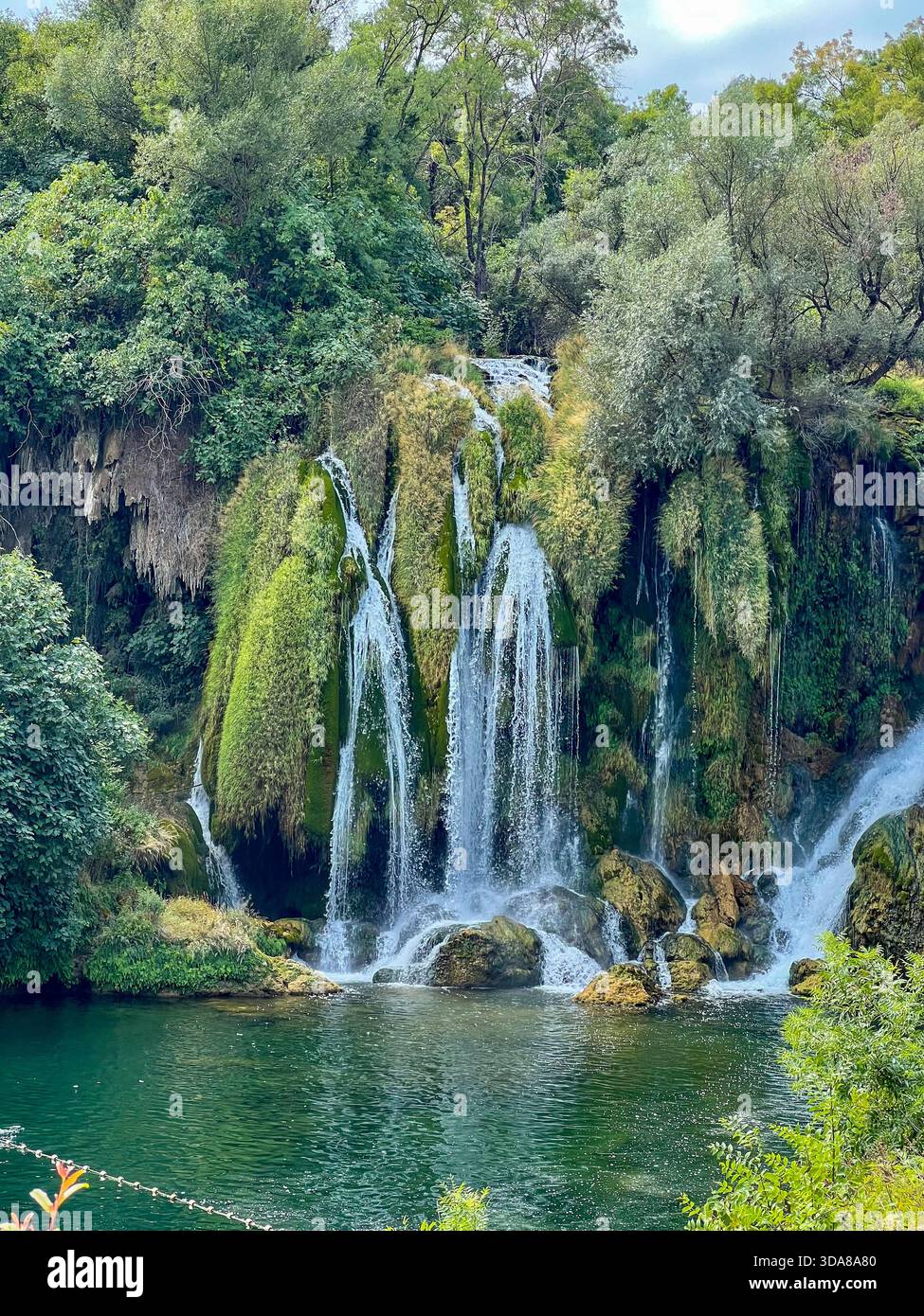 Lebendige Aussicht auf den Kravica Wasserfall in Herzegowina, mit kaskadendem Wasser, moosbedeckten Felsen und dichtem grünen Vegetation, die sich reflektiert - Smartphone-aufgenommenes Stockfoto
