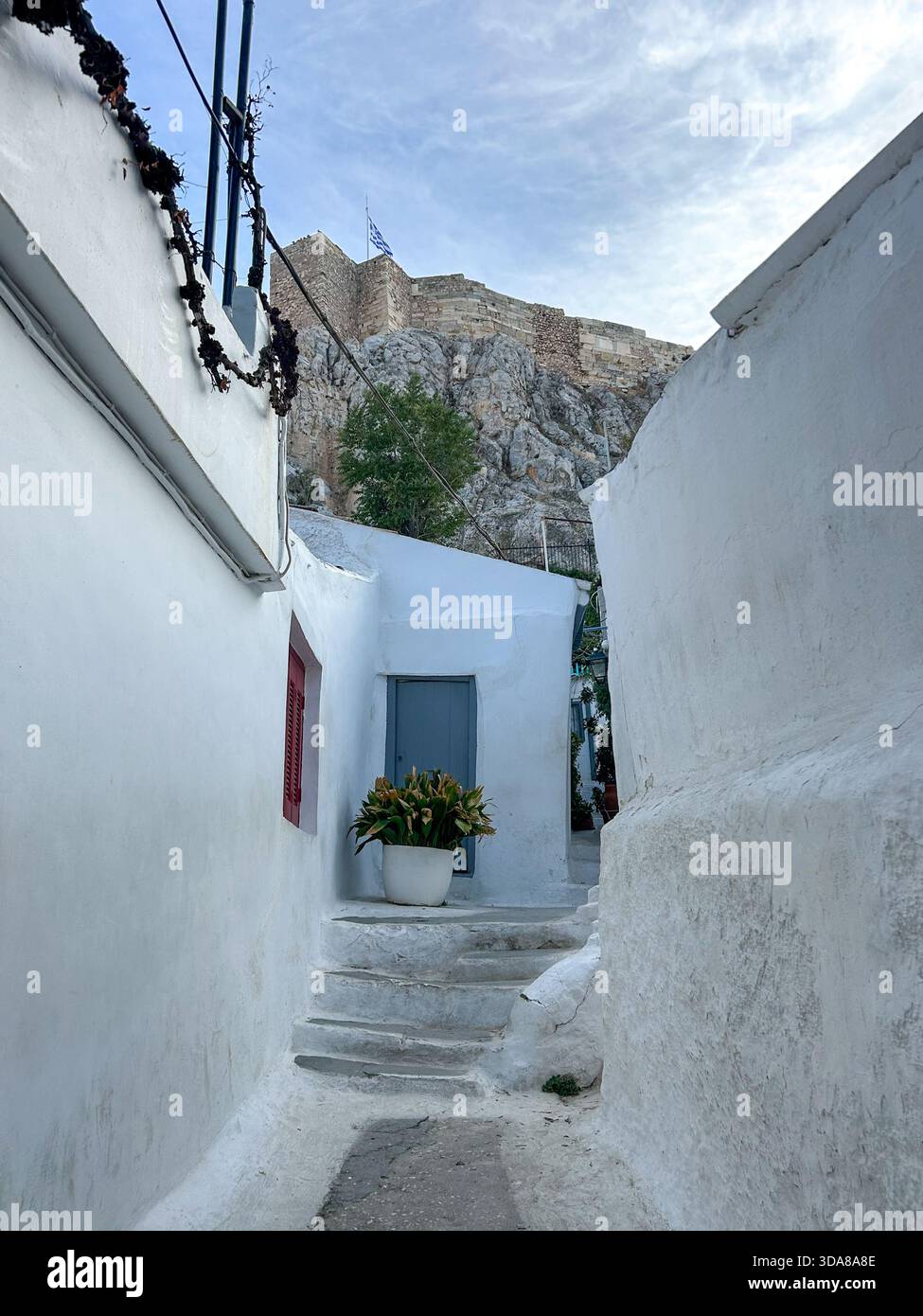 Eine weiß getünchte Gasse in Anafiotika, Athen, mit Häusern im Kykladischen Stil, Treppen und einem Blick auf die Akropolis, die sich über dem historischen Viertel erhebt. - Smartphone-aufgenommenes Stockfoto