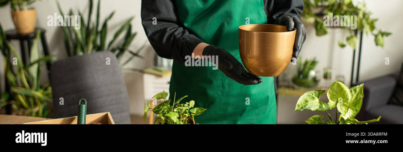 Ein junger asiatischer Blumenhändler hält gekonnt einen goldenen Topf, umgeben von üppigem Grün und Pflanzen. Stockfoto