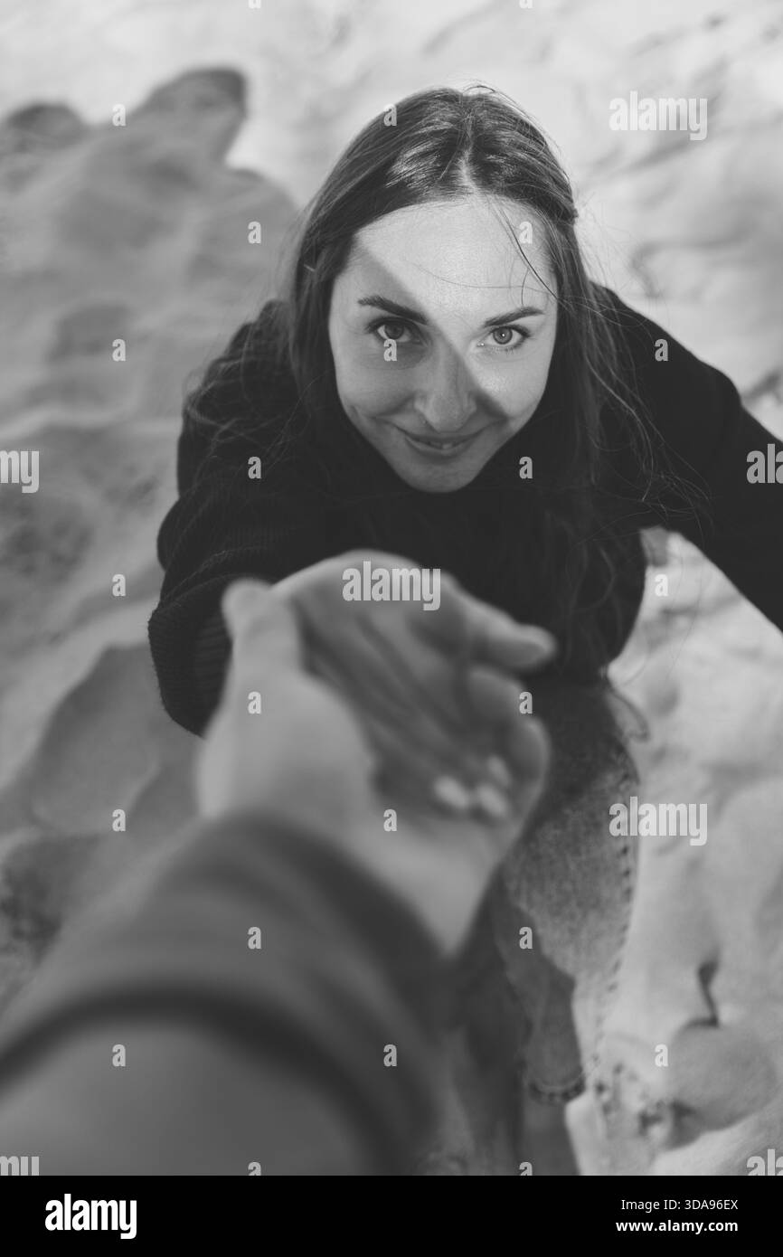 Schwarz-weiße Nahaufnahme einer Frau, die lächelt, während sie am Sandstrand die Hand ausstreckt. Weiches natürliches Licht unterstreicht ihre ausdrucksstarken Augen und ihr freundliches GE Stockfoto
