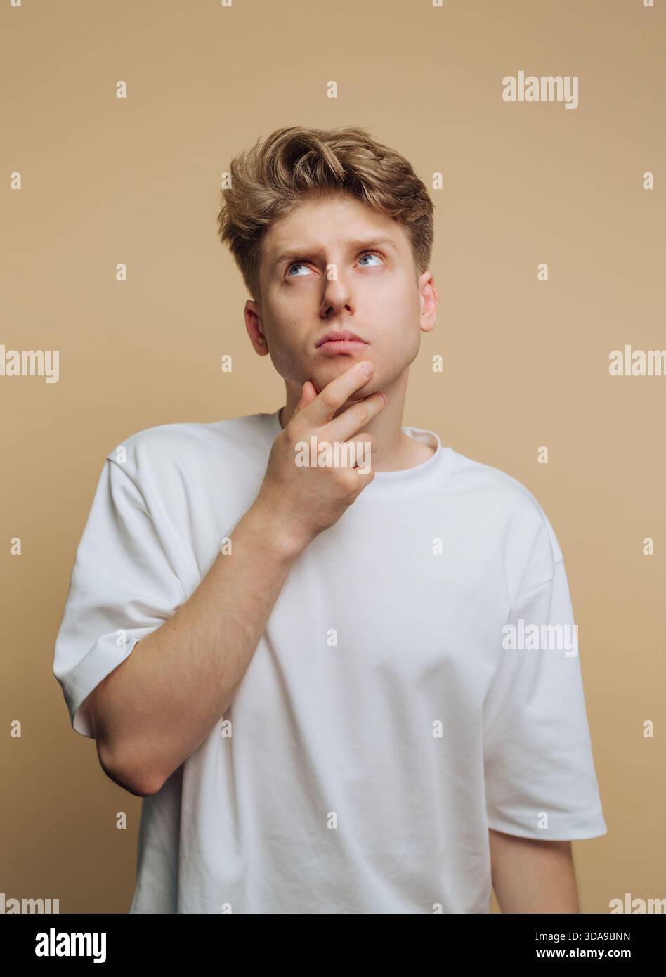 Junger Mann in weißem T-Shirt, der vor beigefarbenem Hintergrund denkt. Stockfoto