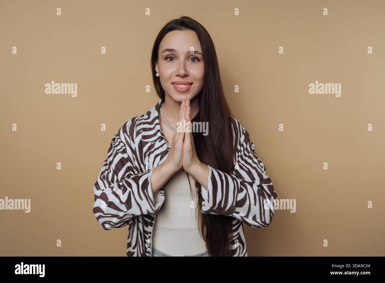 Eine junge Frau steht mit den Händen zusammen in freundlicher Geste. Sie hat lange Haare und trägt eine Jacke mit Zebramuster gegen einen einfarbigen Hintergrund Stockfoto