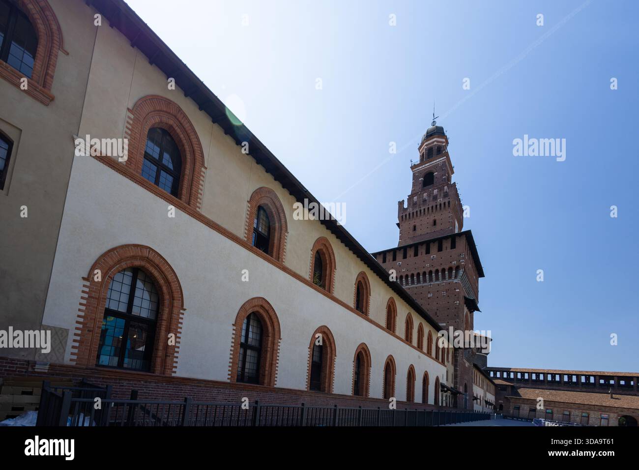 Ein historisches Backsteingebäude mit Rundbogenfenstern und einem hohen Backsteinturm befindet sich entlang einer sonnenbeschienenen Kopfsteinpflasterstraße und schafft eine dramatische Architektur Stockfoto