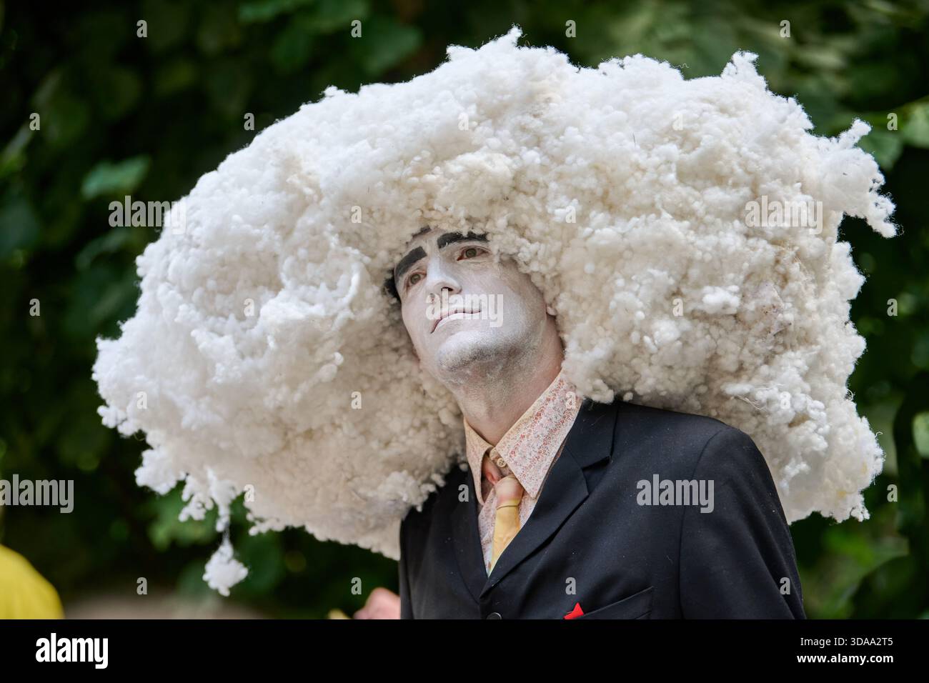 Ein erwachsener männlicher Darsteller mit weißer Gesichtsfarbe trägt einen riesigen, wolkenähnlichen Kopfschmuck, trägt einen dunklen Anzug und Krawatte und posiert nachdenklich vor grünem Laub Stockfoto