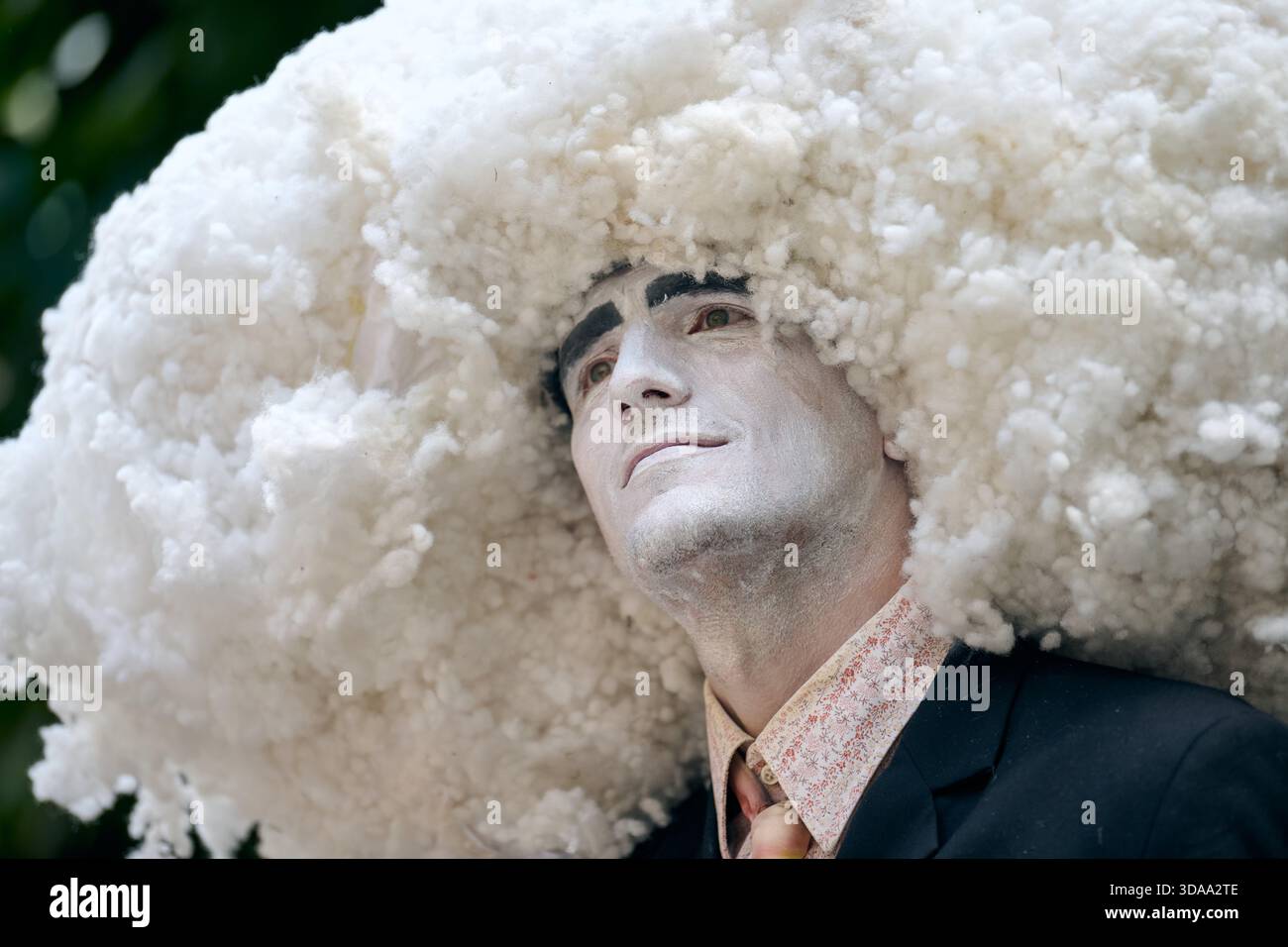Erwachsene männliche Darsteller mit weißer Gesichtsfarbe trägt einen großen, flauschigen, wolkenartigen Kopfschmuck während der Outdoor-Show. Weiches Tageslicht unterstreicht surreale Kostüme Stockfoto