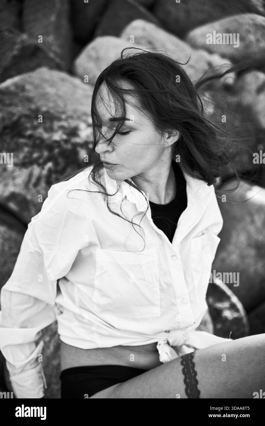 Schwarz-weißes Porträt einer jungen Frau mit dunklem Haar, weißem Hemd und schwarzer Unterwäsche, auf Felsen sitzend, mit im Wind wehenden Haaren, schauend Stockfoto