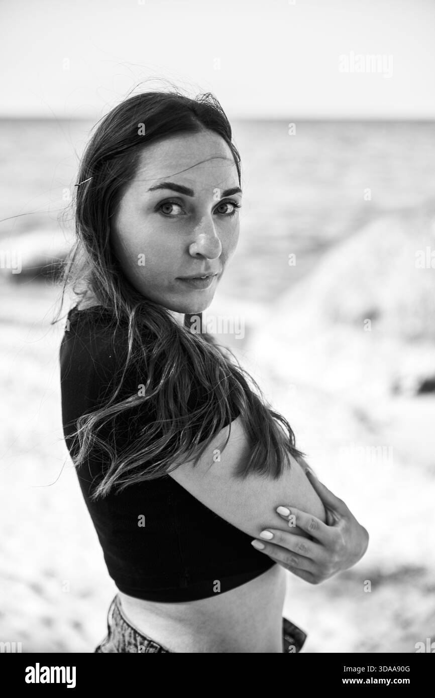 Eine Erwachsene Frau mit langen Haaren steht am Sandy Beach in der Nähe des ruhigen Meeres und blickt leise über ihre Schulter. Schwarzweißbild erfasst kontemplative Stimmung unter Stockfoto