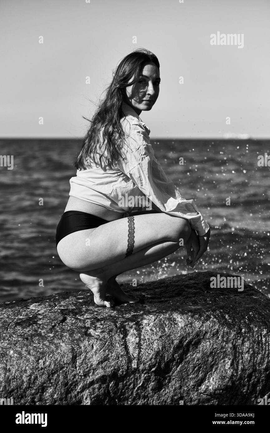 Schwarz-weißes Porträt einer jungen Frau mit langen Haaren, die auf Felsen in der Nähe der Ostseeküste hockt. Ruhiger Meereshorizont und spritzende Wassertröpfchen erzeugen s Stockfoto