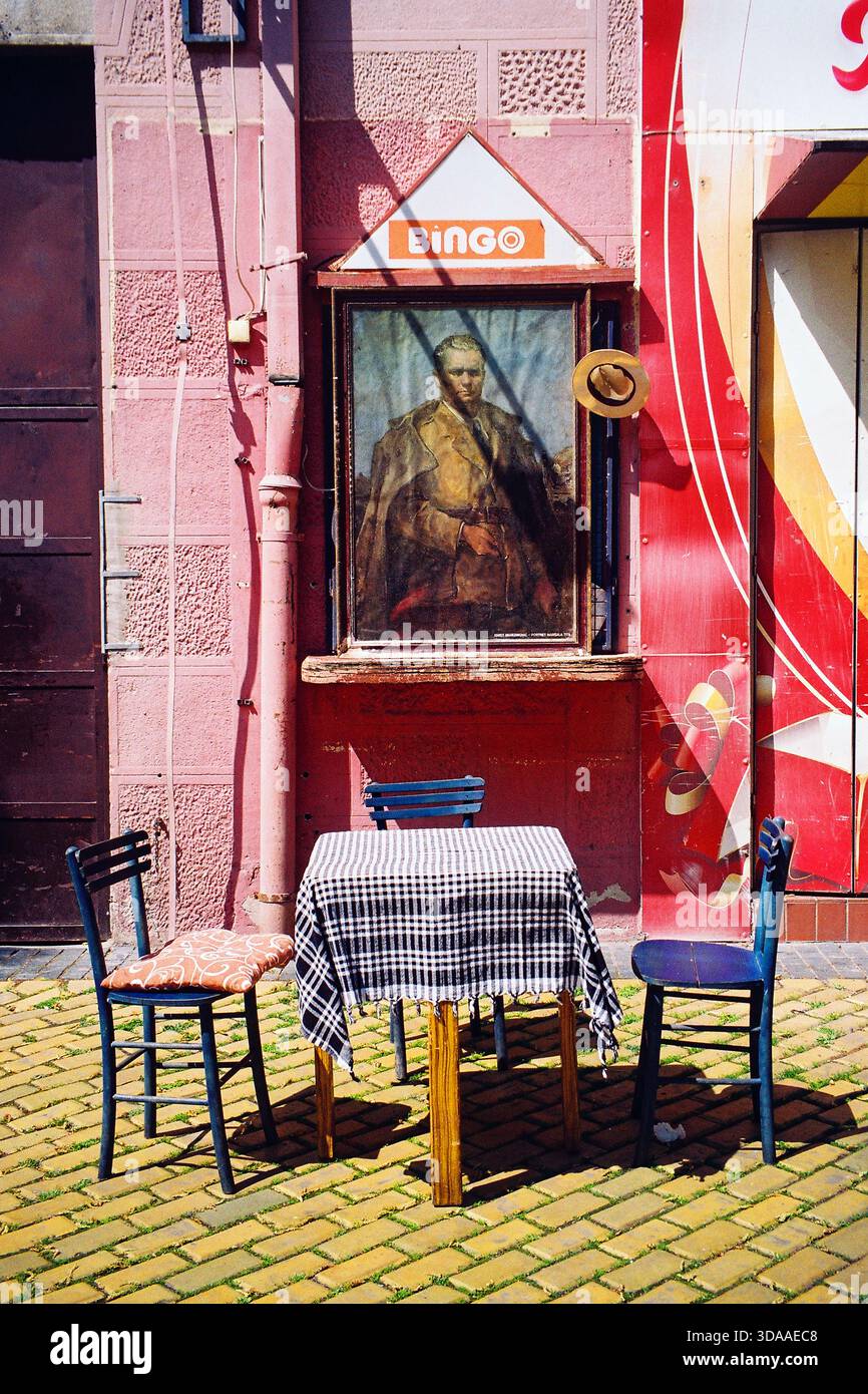 Analoge Aufnahme von drei Stühlen und einem Tisch mit blauem kariertem Tuch auf der Straße, mit einem Tito-Porträt dahinter und einem „Bingo“-Schild darüber, sonnig. Stockfoto