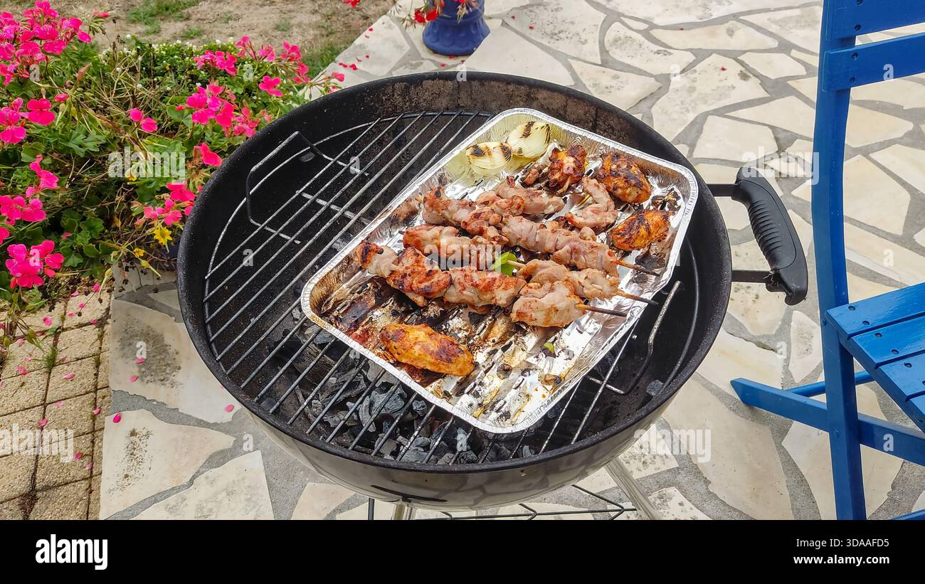 Grill gefüllt mit verschiedenen Fleischsorten auf einer Terrasse umgeben von bunten Blumen. Warmes Außenlicht sorgt für eine entspannte und einladende Atmosphäre. Stockfoto