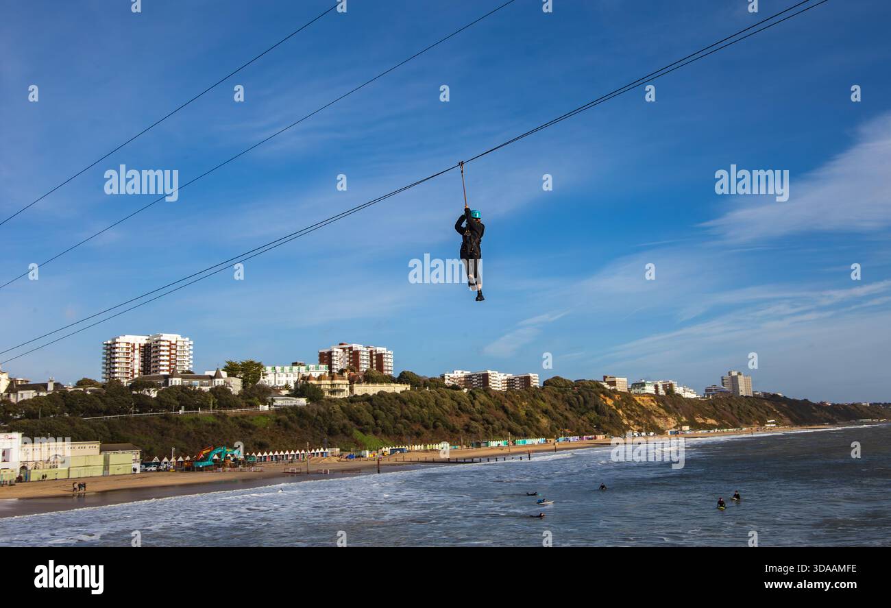 Person Seilrutsche über einem Strand mit Meereswellen und Gebäuden an der städtischen Küste unter einem klaren blauen Himmel. Bournemouth Pier Zip Dezember 2022 Stockfoto