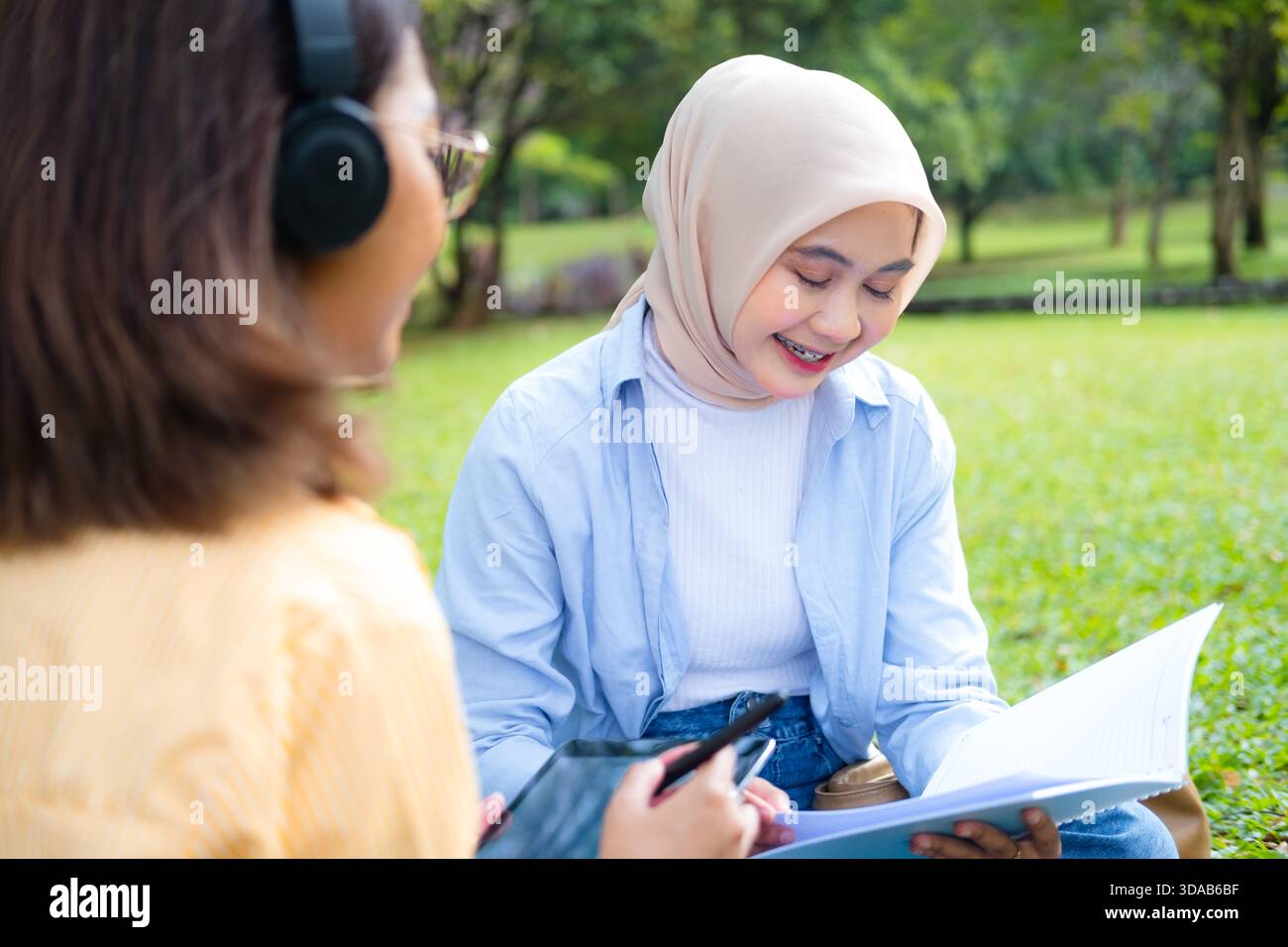 Eine fokussierte asiatische Studentin mit Brille und Kopfhörern hält ein Tablet und einen Stift und denkt während des Lernens im Freien in der Nähe eines modernen Universitätsgebäudes nach Stockfoto