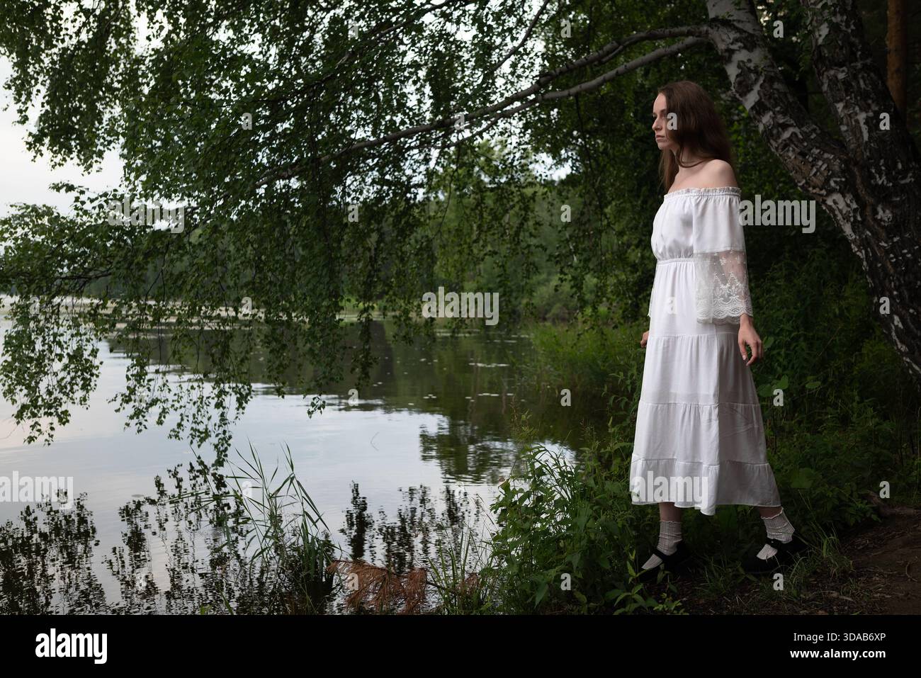 Eleganz am See: Frau in weißem Off-Shoulder Kleid unter Birkenbäumen. Stockfoto
