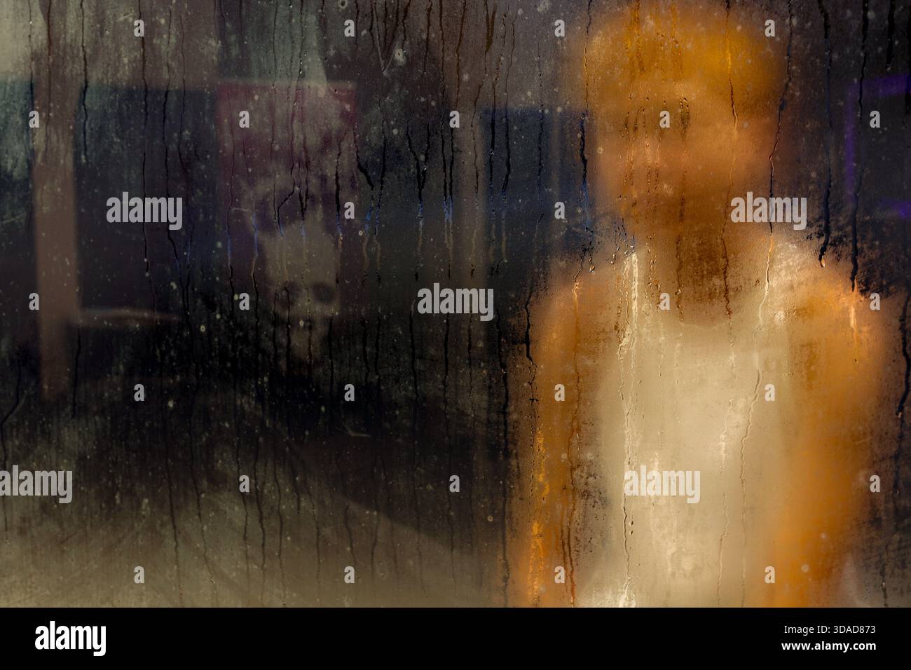 Hinter nassem Glas steht eine abstrakte, verschwommene Figur, in der Wassertropfen und -Streifen eine ausdrucksstarke Textur bilden Stockfoto