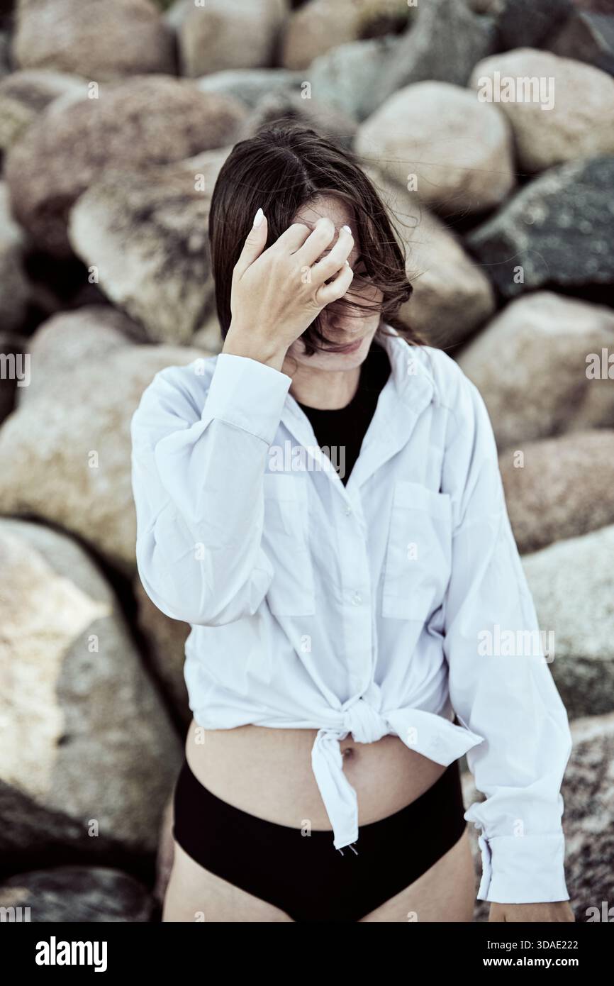 Die junge Frau, die ein gebundenes weißes Hemd und eine schwarze Bikinihose trägt, steht vor dem Hintergrund großer Strandsteine. Natürliches Licht hebt offen, introspe hervor Stockfoto