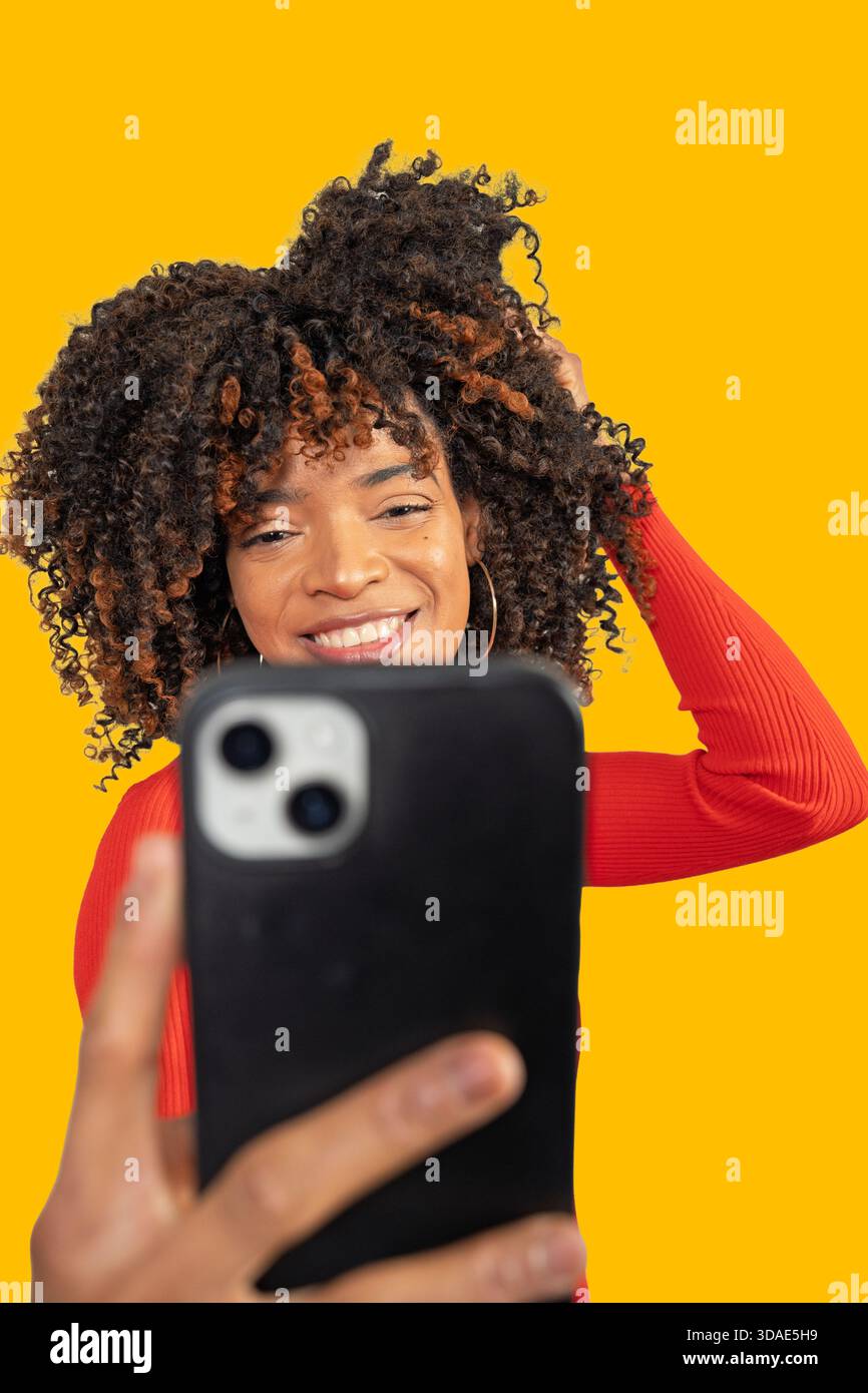 Glückliche junge Frau mit lockigem Haar, die Videos filmt oder Selfie vor gelbem Hintergrund macht Stockfoto