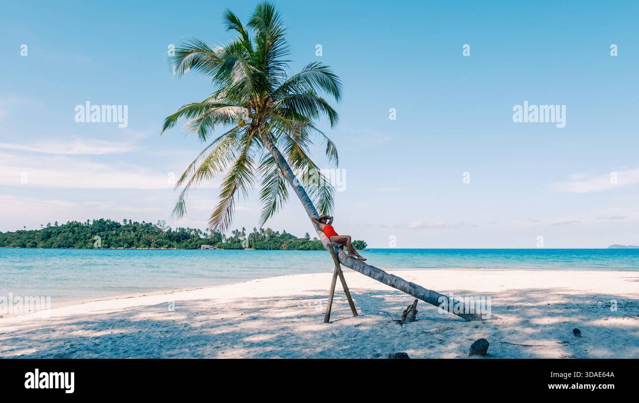 Eine Asiatin genießt einen ruhigen Moment, um sich an einer hohen Palme am weißen Sandstrand von Koh Kood Island zu entspannen. Das türkisfarbene Wasser glitzert unter einem klaren blauen Himmel und schafft ein tropisches Paradies. || Modell freigegeben Stockfoto