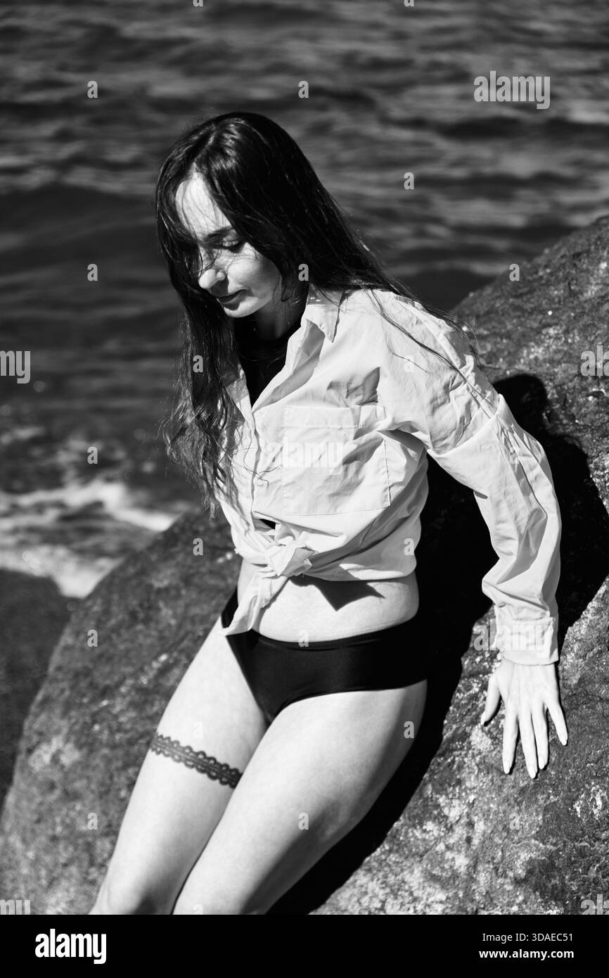 Schwarz-weißes Porträt einer jungen Frau mit dunklem Haar in weißem Hemd und schwarzem Unterteil, auf Felsen in der Nähe der Ostsee. Moody vor dem Meer Stockfoto