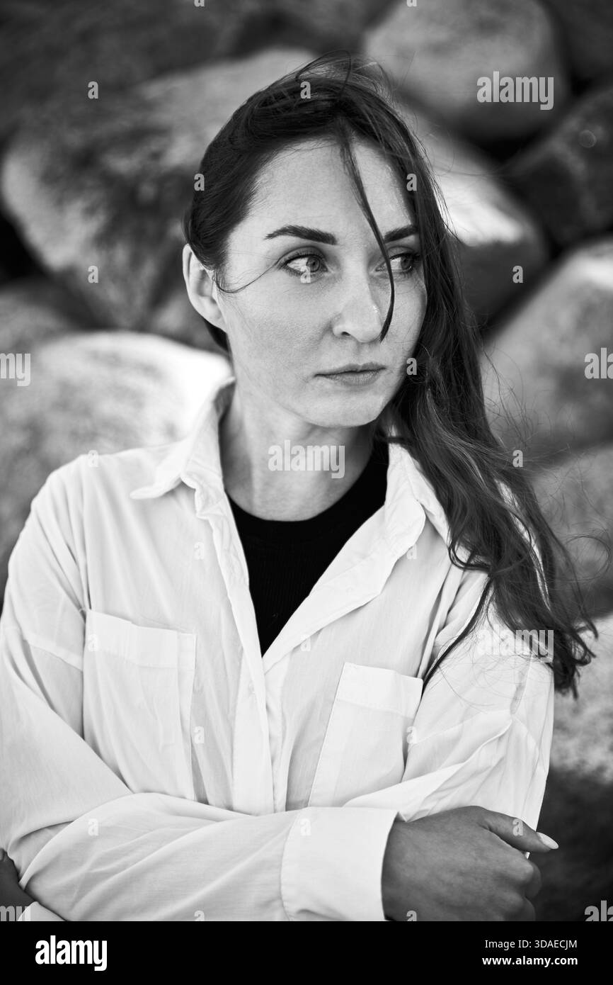 Schwarz-weißes Porträt einer jungen Frau mit langen dunklen Haaren, die seitlich gucken, mit weißem Hemd über schwarzem Oberteil. Natürliche felsige Umgebung und weiches li Stockfoto
