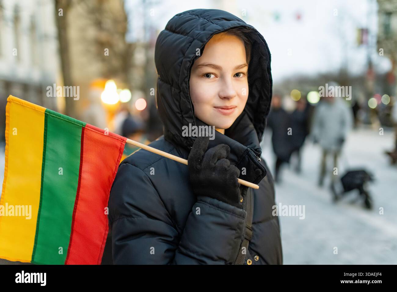 Mädchen in einem schwarzen Wintermantel, das die litauische Flagge hält, mit einem leisen Lächeln während der Feier des Unabhängigkeitstages. Ein stolzer, friedlicher Moment im Herzen Stockfoto
