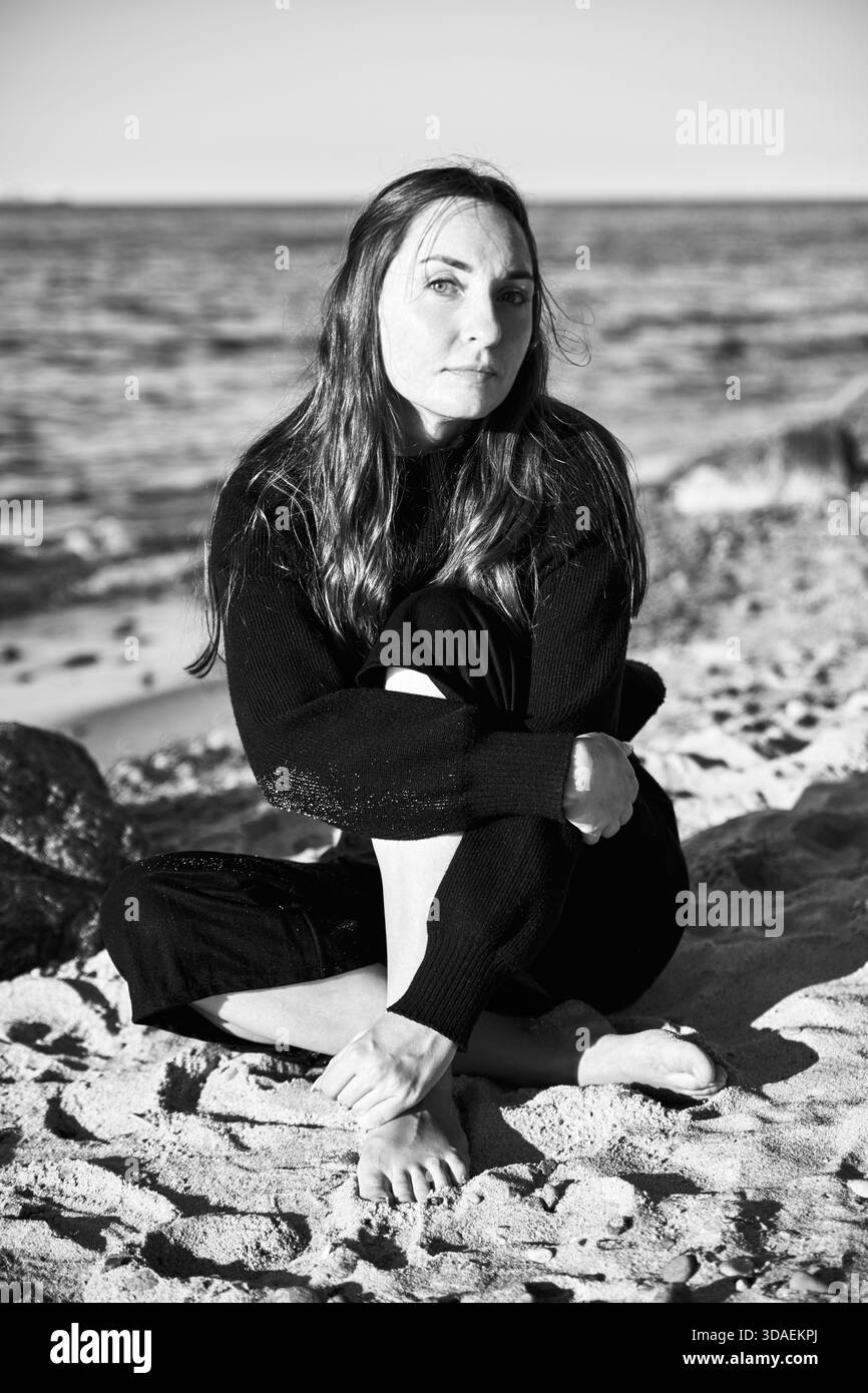 Porträt einer jungen Frau mit langen Haaren barfuß am Sandstrand an der Ostsee. Natürliches Licht hebt den ruhigen Hintergrund des Meeres und die ruhige Ausdrucksweise hervor Stockfoto