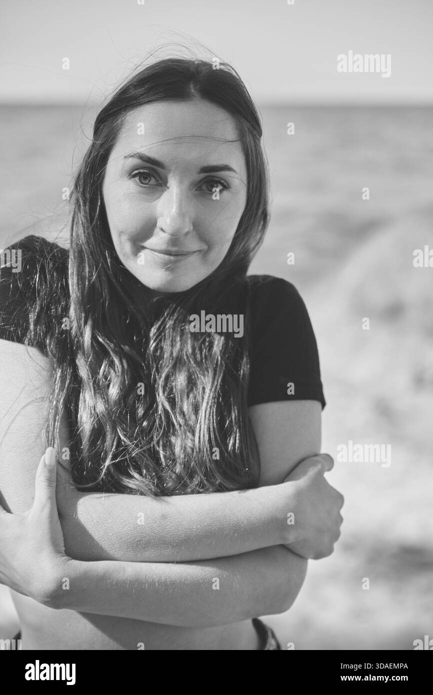 Nahaufnahme eines Porträts einer jungen Frau mit langen dunklen Haaren, die sich in der Nähe des Strandes umhüllt. Sanftes Tageslicht und einfarbige Farbtöne sorgen für intime, besinnliche Atmosphäre Stockfoto