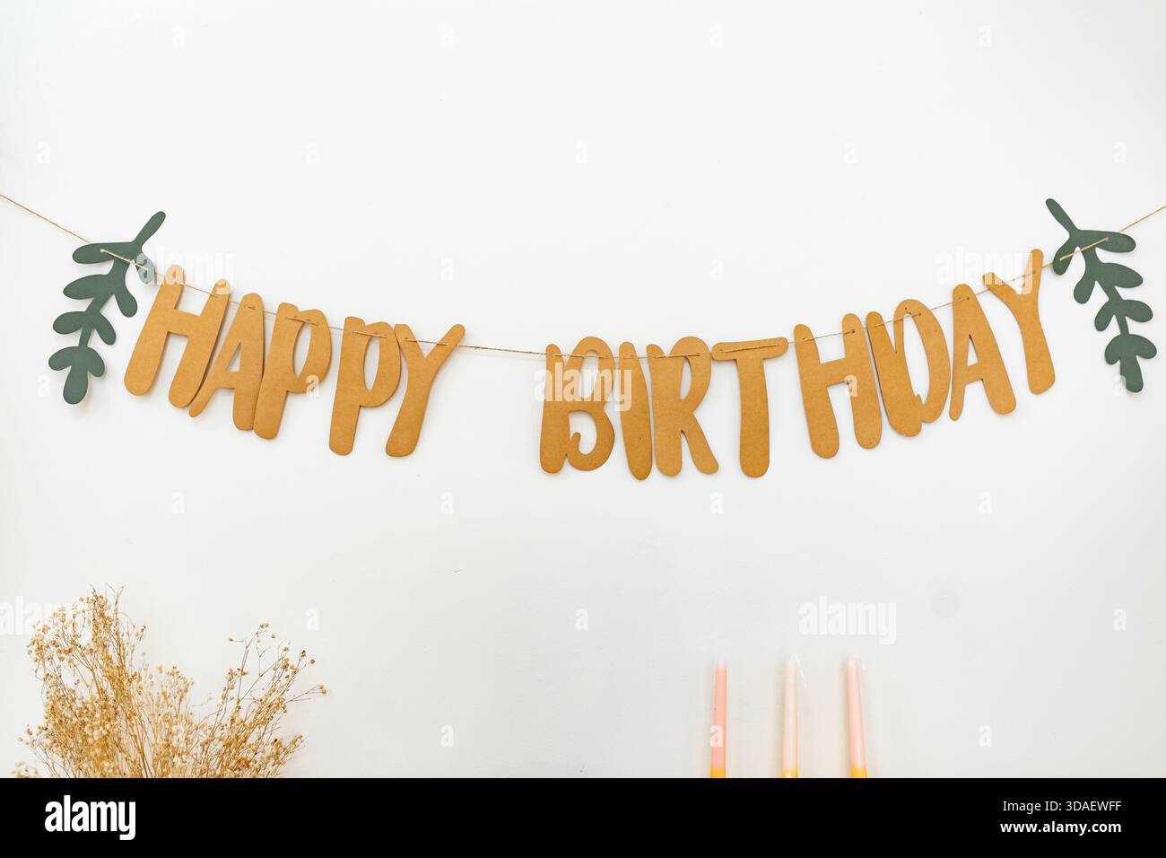 Ein festliches Happy Birthday-Banner aus braunen Buchstaben mit grünen grünen Blattakzenten hängt an einer weißen Wand. Unten kommen Pastellkerzen und getrocknete Blumen hinzu Stockfoto