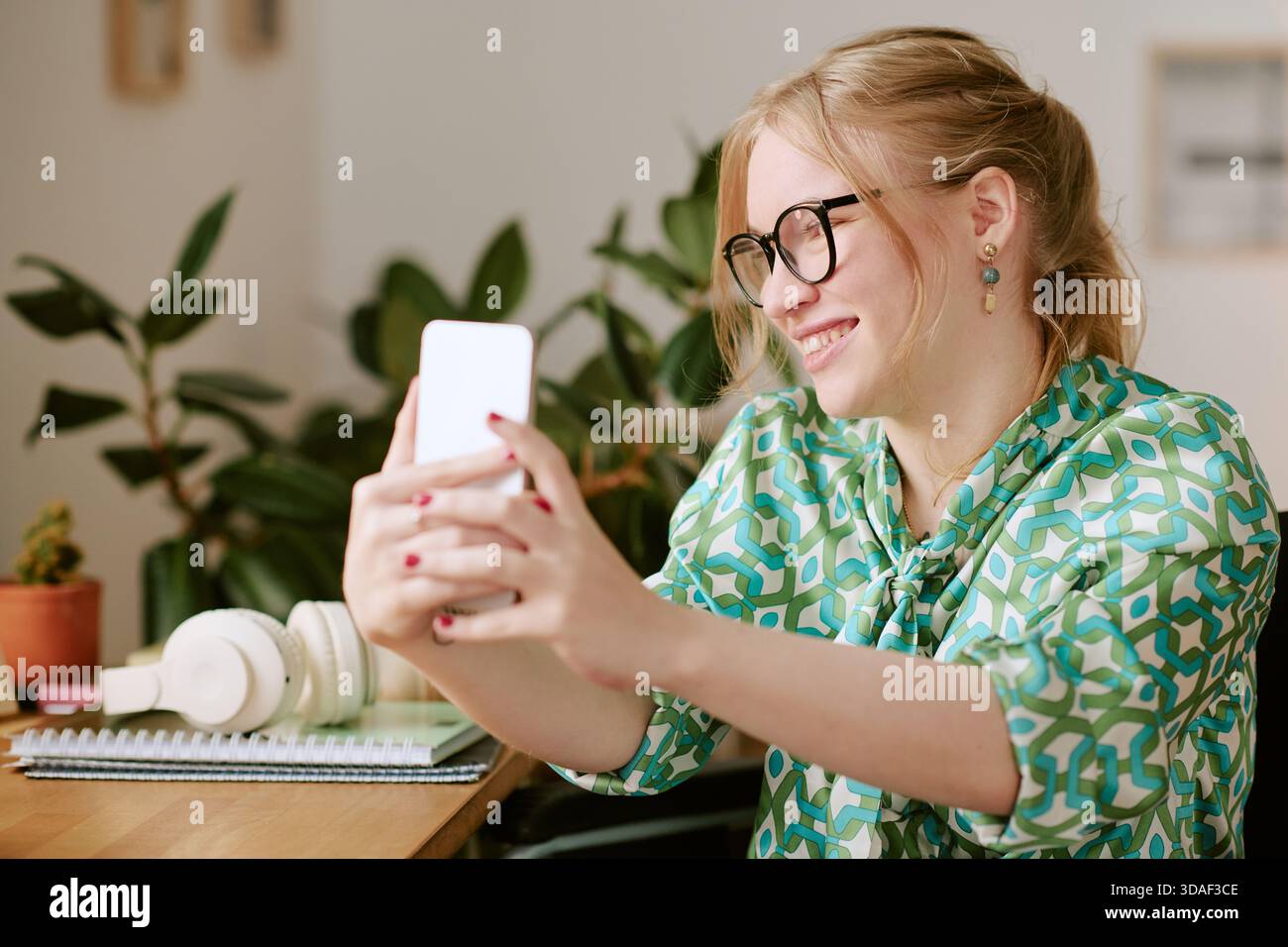 Junge Frau lächelt, während sie Selfie mit Smartphone in Innenräumen nimmt Stockfoto
