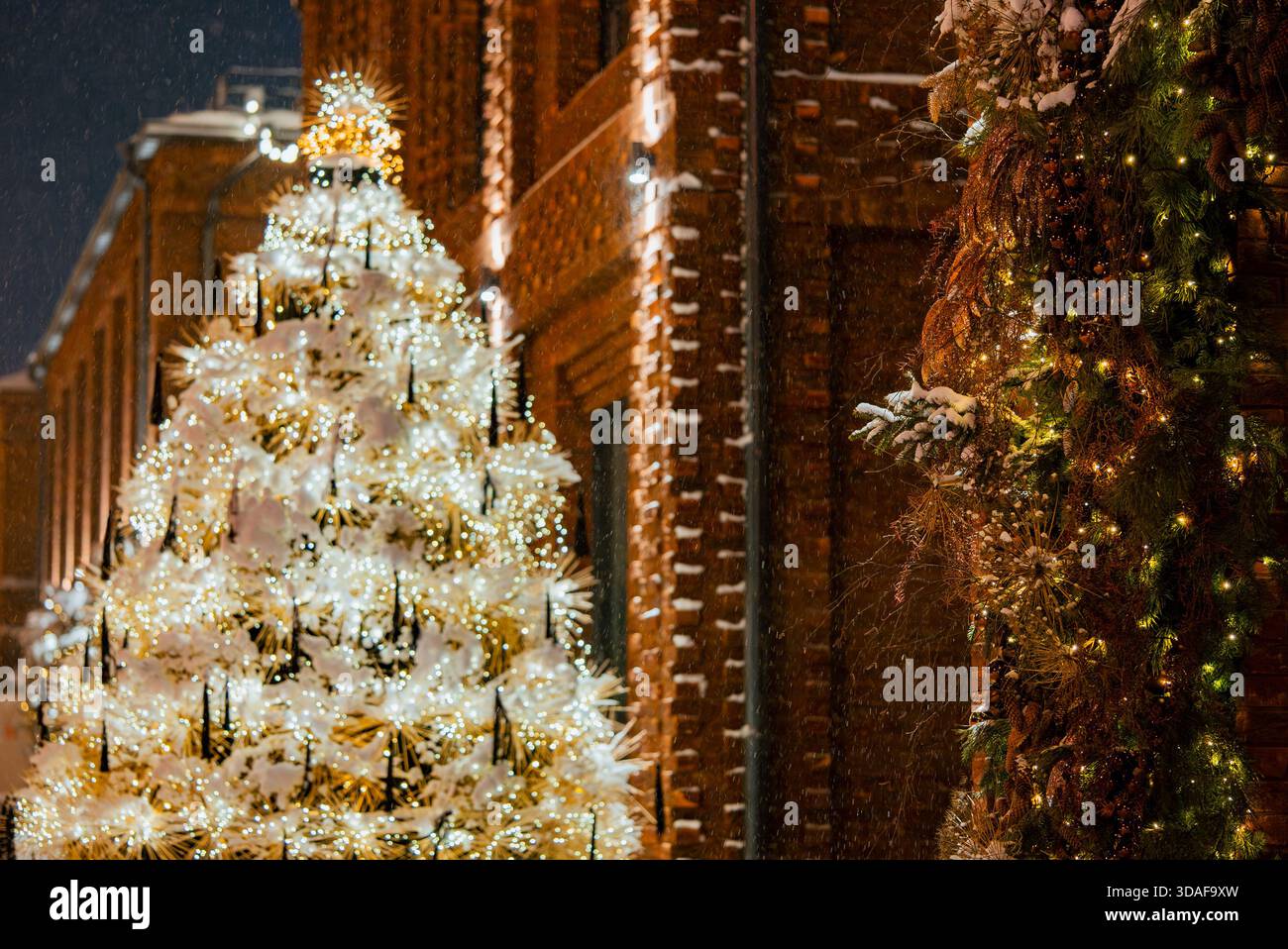 Der hell beleuchtete weihnachtsbaum mit warmweißen Lichtern und Schnee steht vor einem Backsteingebäude und schafft eine festliche Winterszene an einem schneebedeckten Feiertagsabend Stockfoto