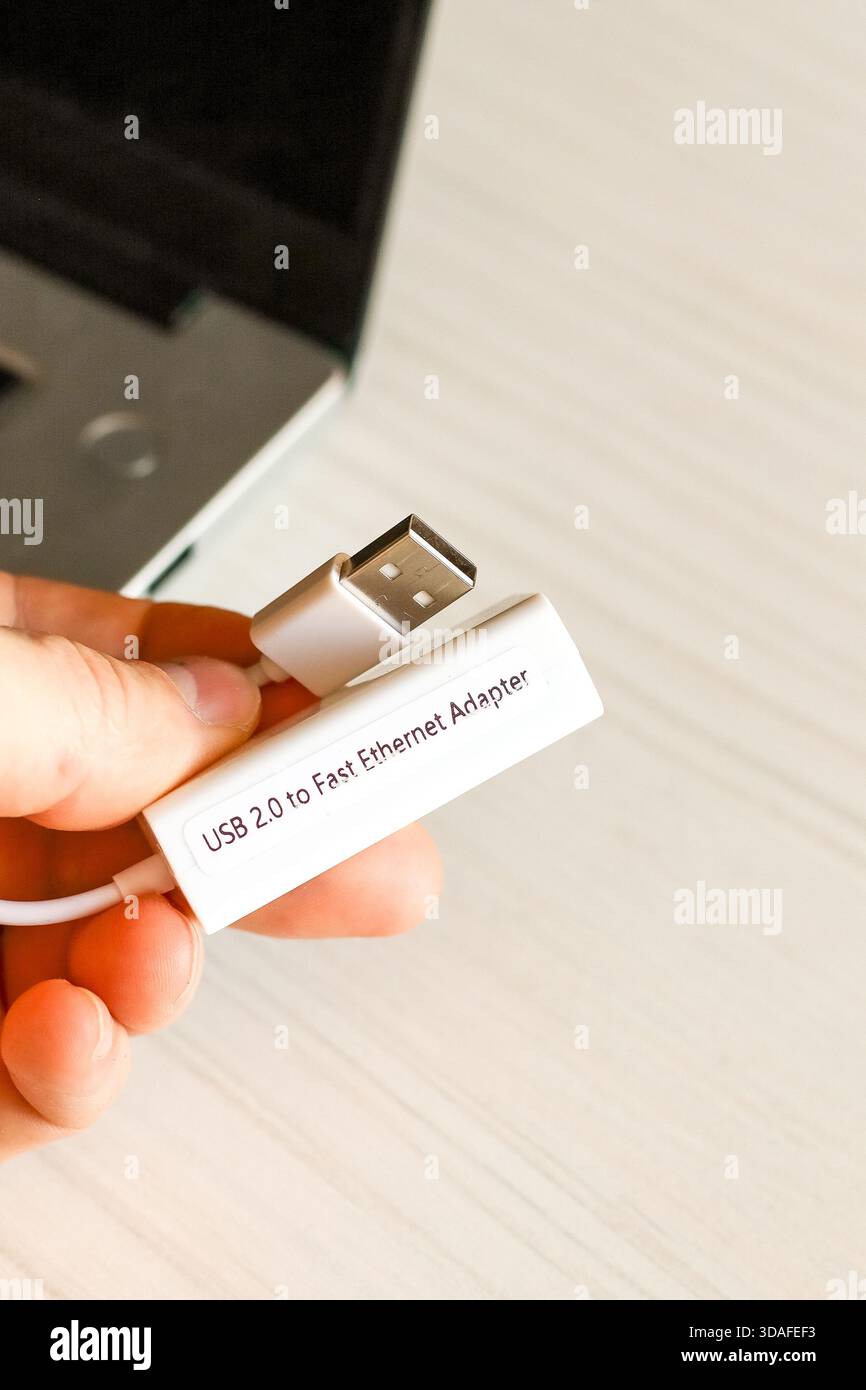 Die Hand des Einzelnen präsentiert ein weißes USB 2,0-auf-Fast-Ethernet-Adapterkabel, das mit einem silbernen Laptop auf einem Schreibtisch verbunden ist, ideal für ein Upgrade auf eine kabelgebundene Verbindung Stockfoto