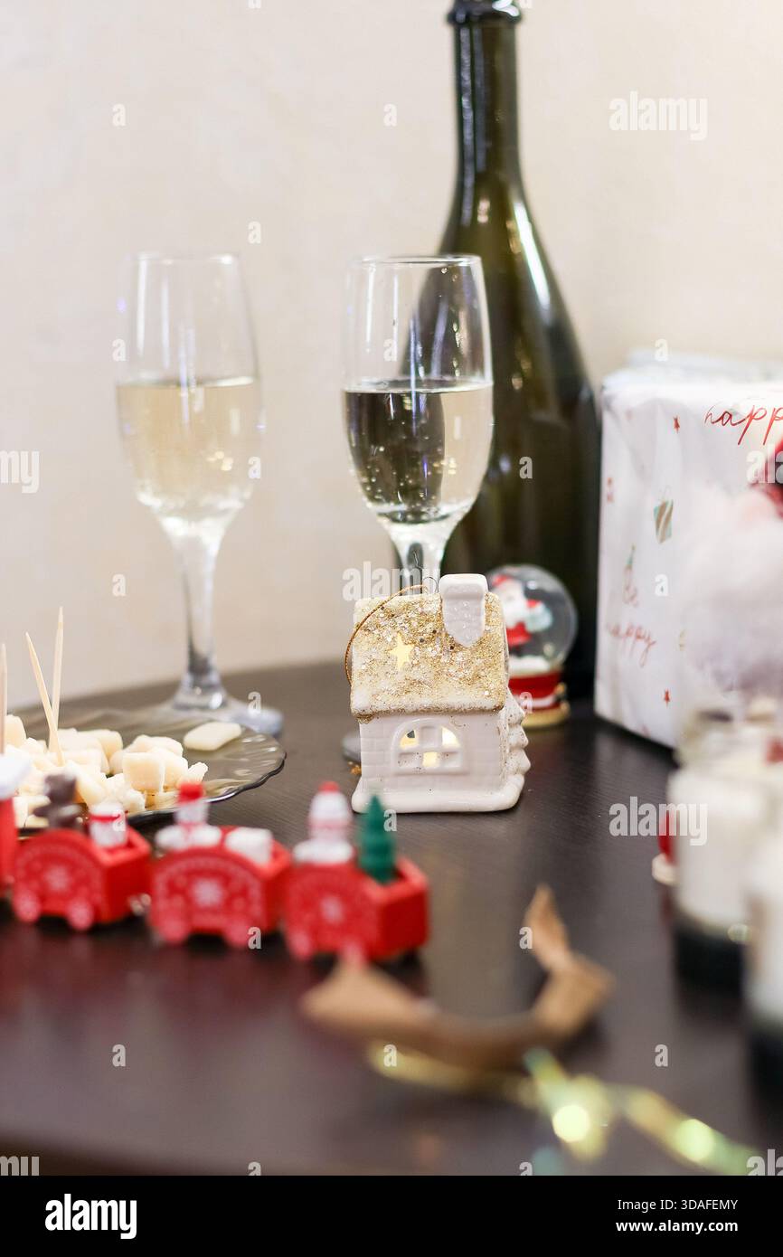 Intimer Blick auf Sekt in Flöten neben der Flasche, mit Käsesnacks, beleuchteter Kerze, kleinem Hausschmuck und Spielzeugzug auf dunkler Oberfläche. Stockfoto