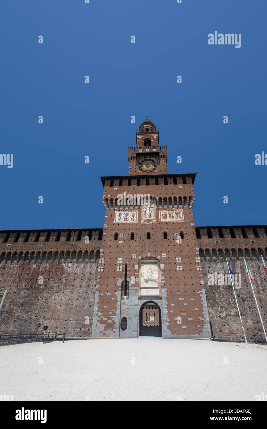 Eindrucksvolle mittelalterliche Festungsfassade mit einem zentralen Glockenturm, dekorativen Mosaiken und Steinreliefs. Breiter, sonnendurchfluteter Innenhof und Fahnenrahmen Stockfoto