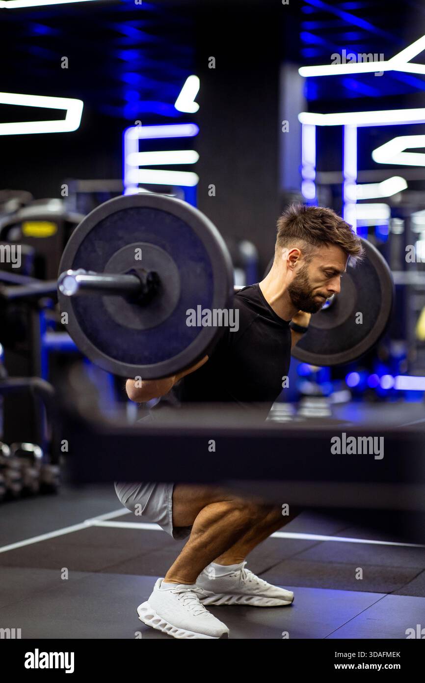 Ein Mann in einem schwarzen Hemd und Shorts konzentriert sich auf seine Form, während er Hocken mit einer Langhantel macht. Der Fitnessraum verfügt über moderne Geräte und farbenfrohe Lichter Stockfoto