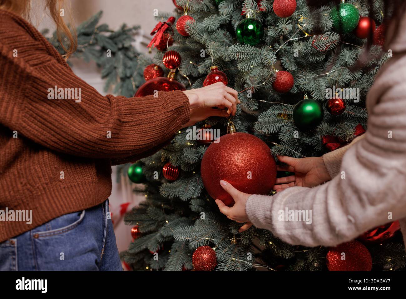 Hände von zwei Frauen, die in einem gemütlichen festlichen Wohnzimmer eine große rote Glitzerkugel an einem geschmückten Weihnachtsbaum hängen. Stockfoto