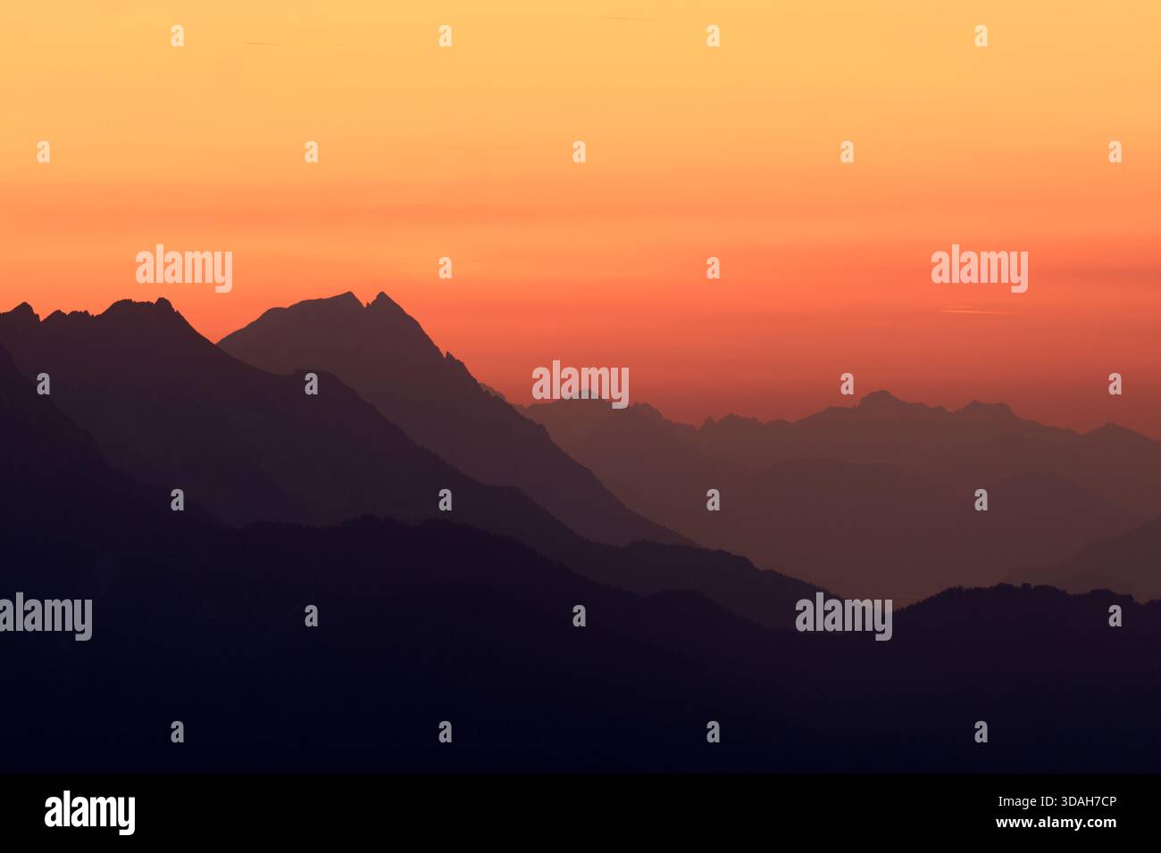 Eine ruhige Berglandschaft bei Sonnenuntergang, mit silbrigen Gipfeln, mehrschichtigen Bergrücken und einem farbenfrohen orangen Himmel, die einen ruhigen und Panoramablick schaffen. Stockfoto