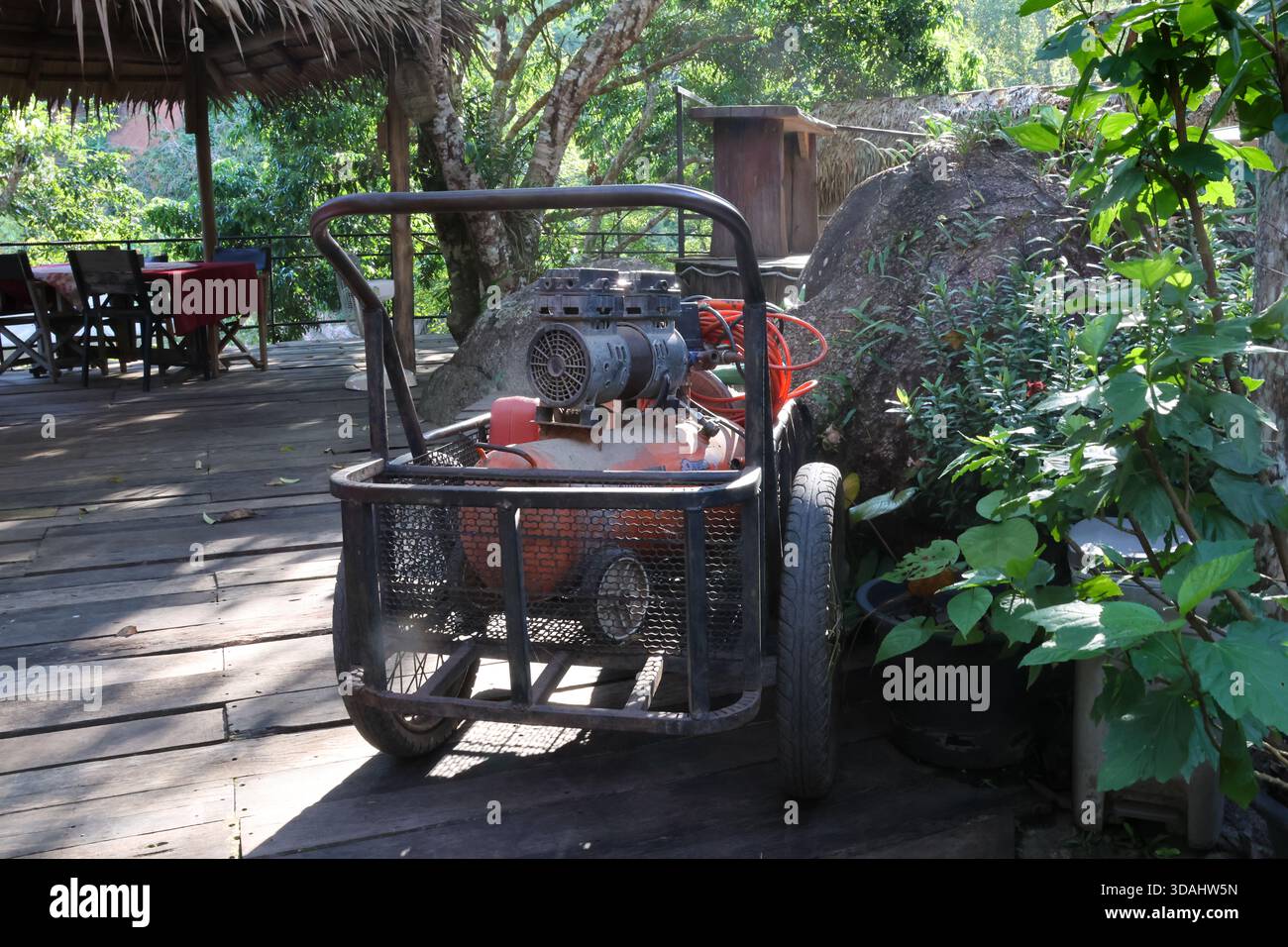 Alte, rustikale Wasserpumpmaschine auf dem Wagen, die auf Holzdeck im üppigen grünen Dschungel ruht. Außenausstattung im tropischen Garten für einfaches ländliches Leben Stockfoto