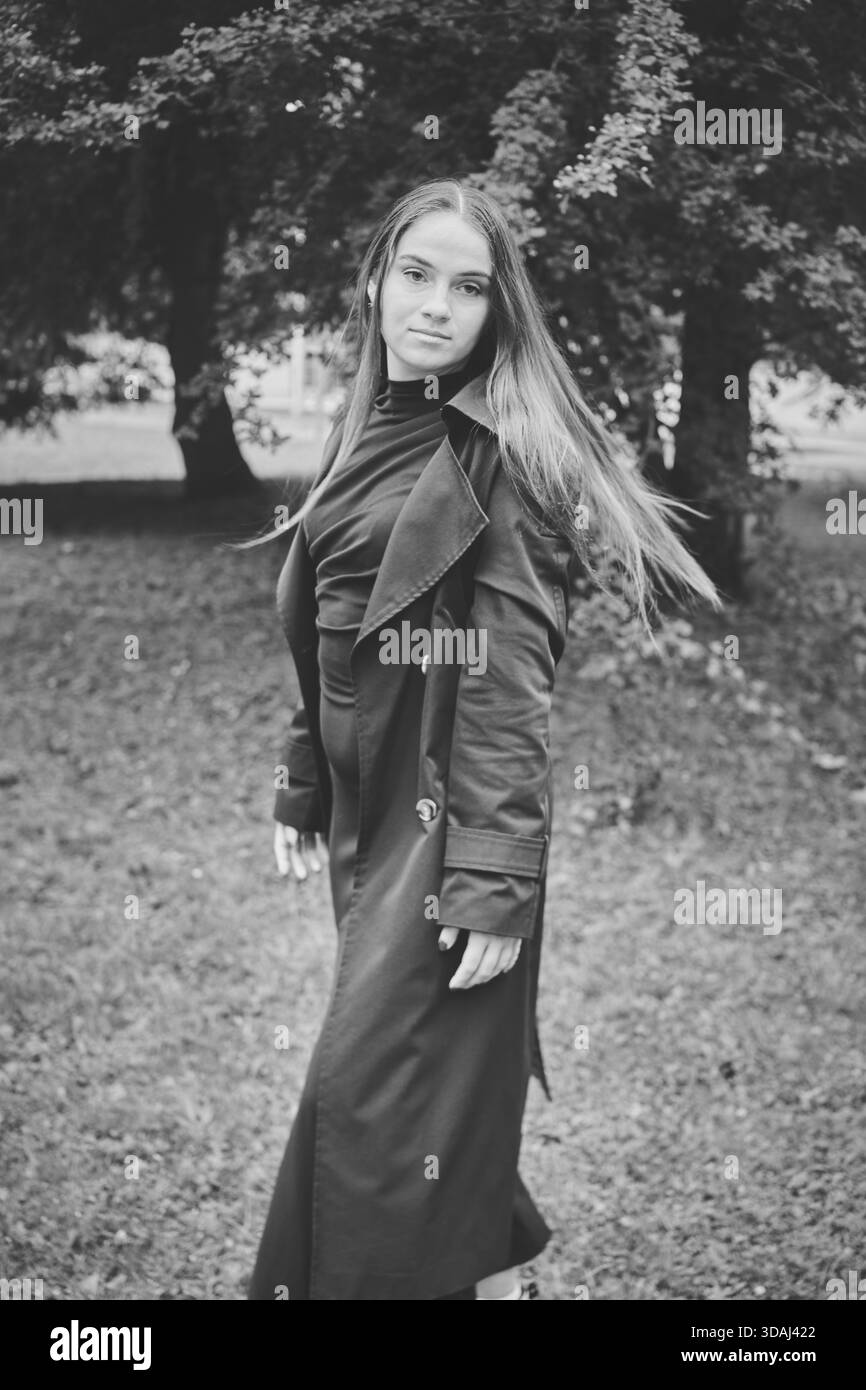 Junge Frau mit langen Haaren und Trenchcoat steht auf Gras unter einem großen Baum und blickt ruhig in die Kamera bei weichem Tageslicht. Hintergrund zeigt Unschärfe Stockfoto