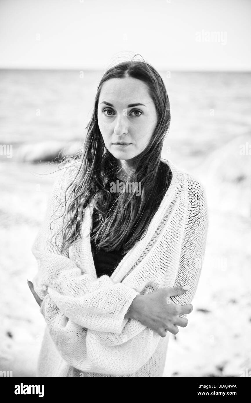 Schwarz-weißes Porträt einer jungen Frau, die am Ostseestrand steht, in einen Strickpullover mit ruhigem, besinnlichem Ausdruck gewickelt. Verschwommenes Meer und Stockfoto