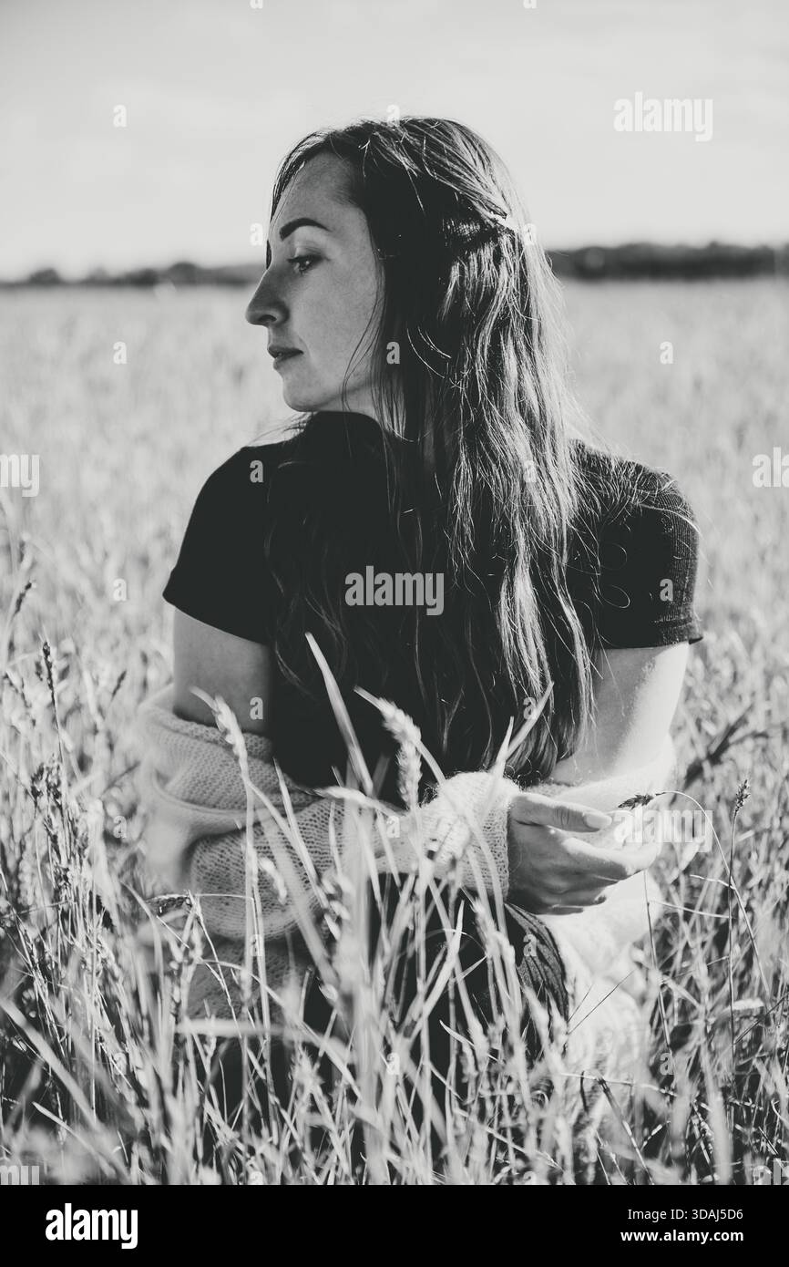 Profilansicht einer jungen Frau mit langen Haaren, die auf freiem Feld zwischen hohen Roggenstielen sitzt. Schwarzweiß-Foto macht ruhige, introspektive Stimmung mit Stockfoto
