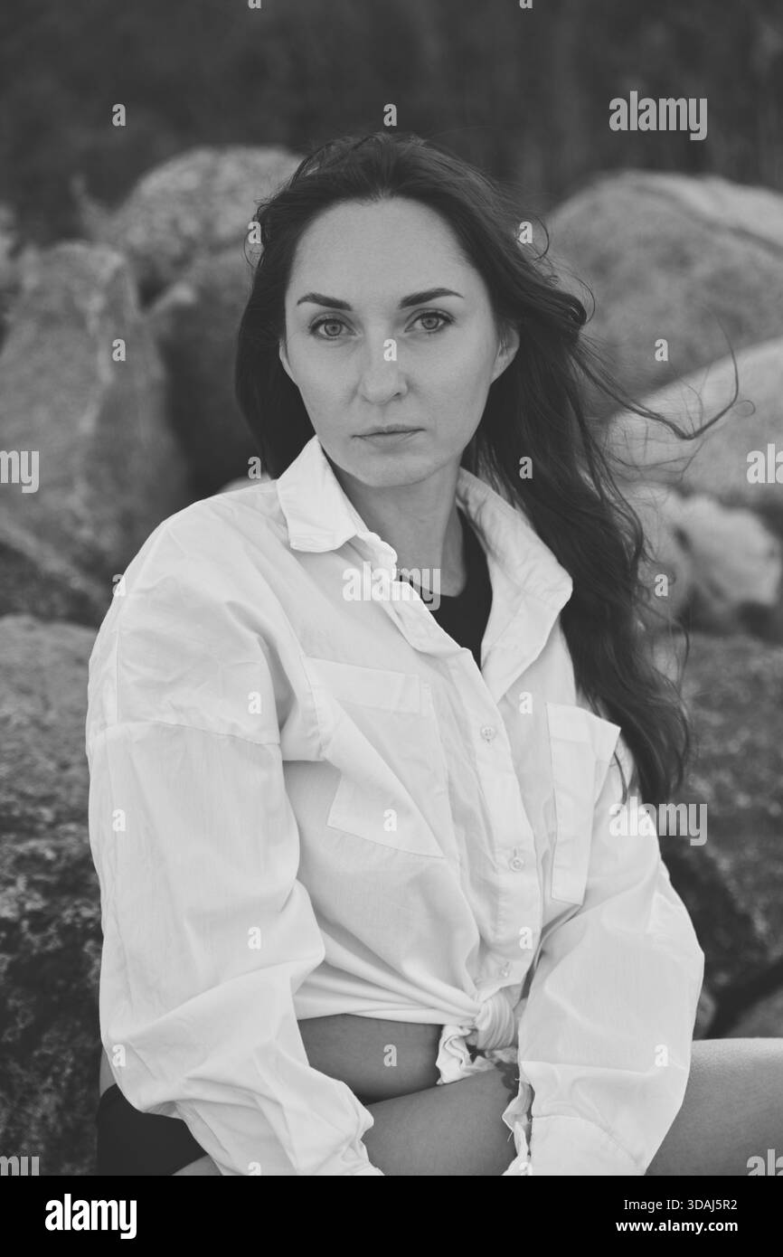 Junge Frau mit langen dunklen Haaren posiert für besinnliches Schwarz-weiß-Porträt draußen von großen Felsen. Natürliches Licht und sanfter Wind tragen dazu bei Stockfoto