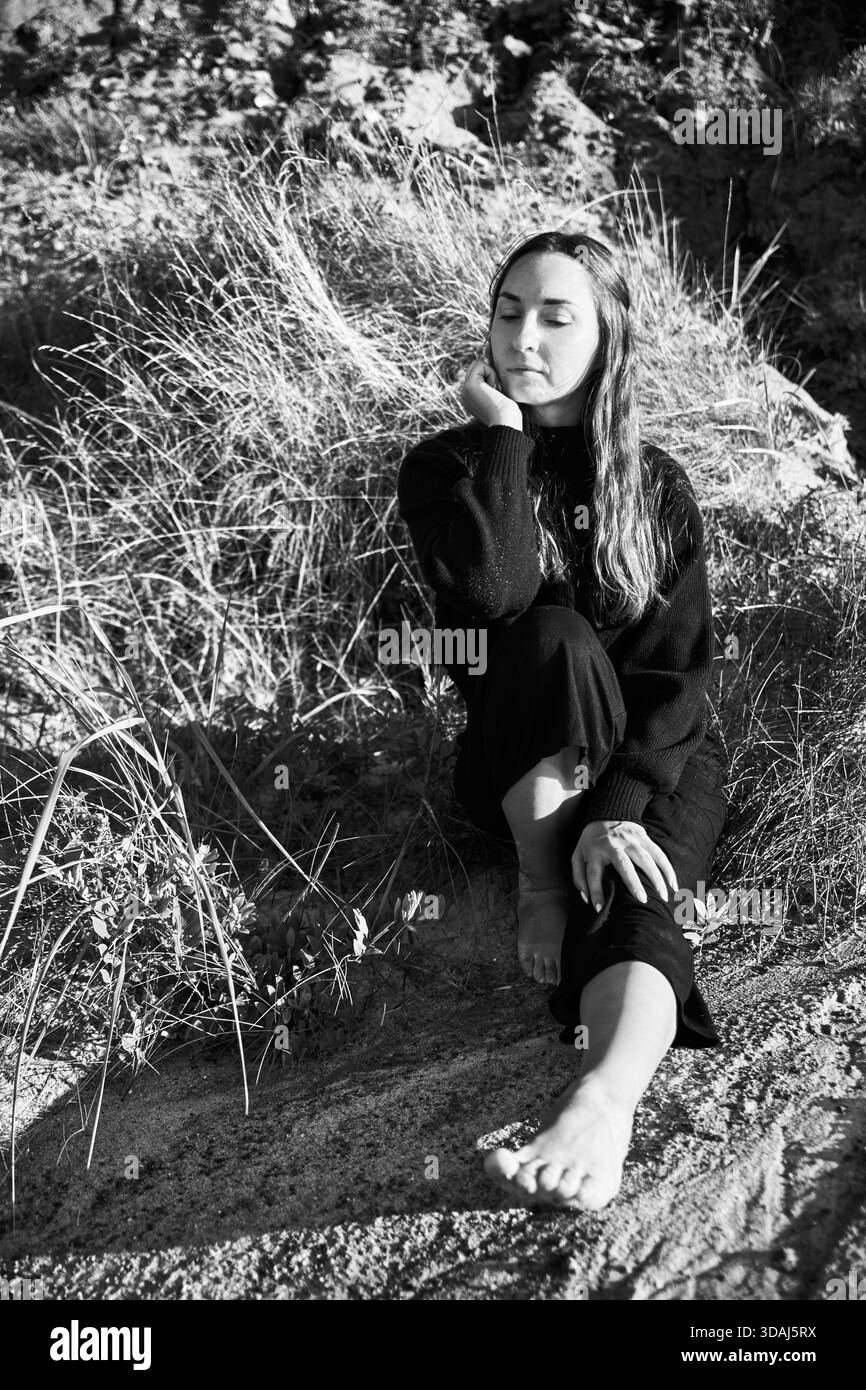 Junge Frau mit langen Haaren in dunkler Kleidung sitzt barfuß auf sonnendurchflutetem Erdboden in der Nähe von hohem Gras. Das Schwarzweiß-Porträt fängt Ruhe und Ruhe ein Stockfoto
