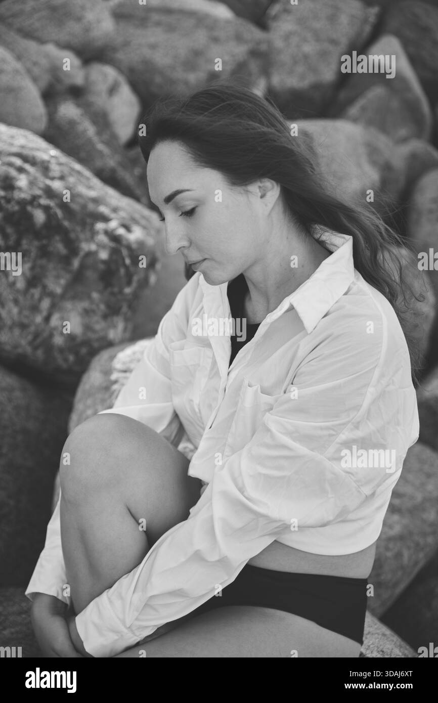 Schwarz-weißes Profilporträt einer jungen Frau mit langen dunklen Haaren, die besinnlich auf großen Felsen sitzt und ein weißes Hemd trägt. Natürliches Licht, Emph Stockfoto