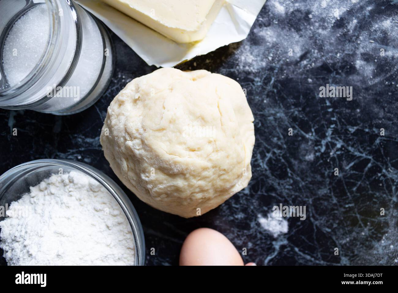 Frischer, hausgemachter Teig in der Mitte einer dunklen Küchenarbeitsfläche, umgeben von Backzutaten wie Eiern, Mehl, Butter und Zucker. Flo Stockfoto