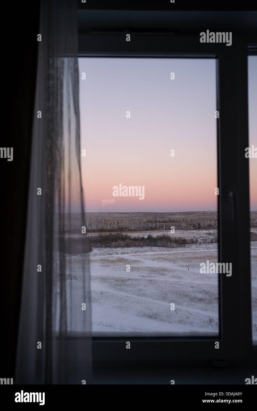 Die Landschaft des Winters bei Sonnenaufgang wird durch einen Fensterrahmen mit einem transparenten Vorhang betrachtet und bietet einen ruhigen und introspektiven Blick auf ein schneebedecktes Feld und einen weit entfernten Wald Stockfoto