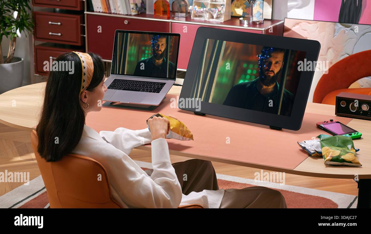 Junge Frau, die sich Filme auf einem großen Tablet-Bildschirm ansieht, während sie in einem gemütlichen Wohnzimmer Chips isst Stockfoto